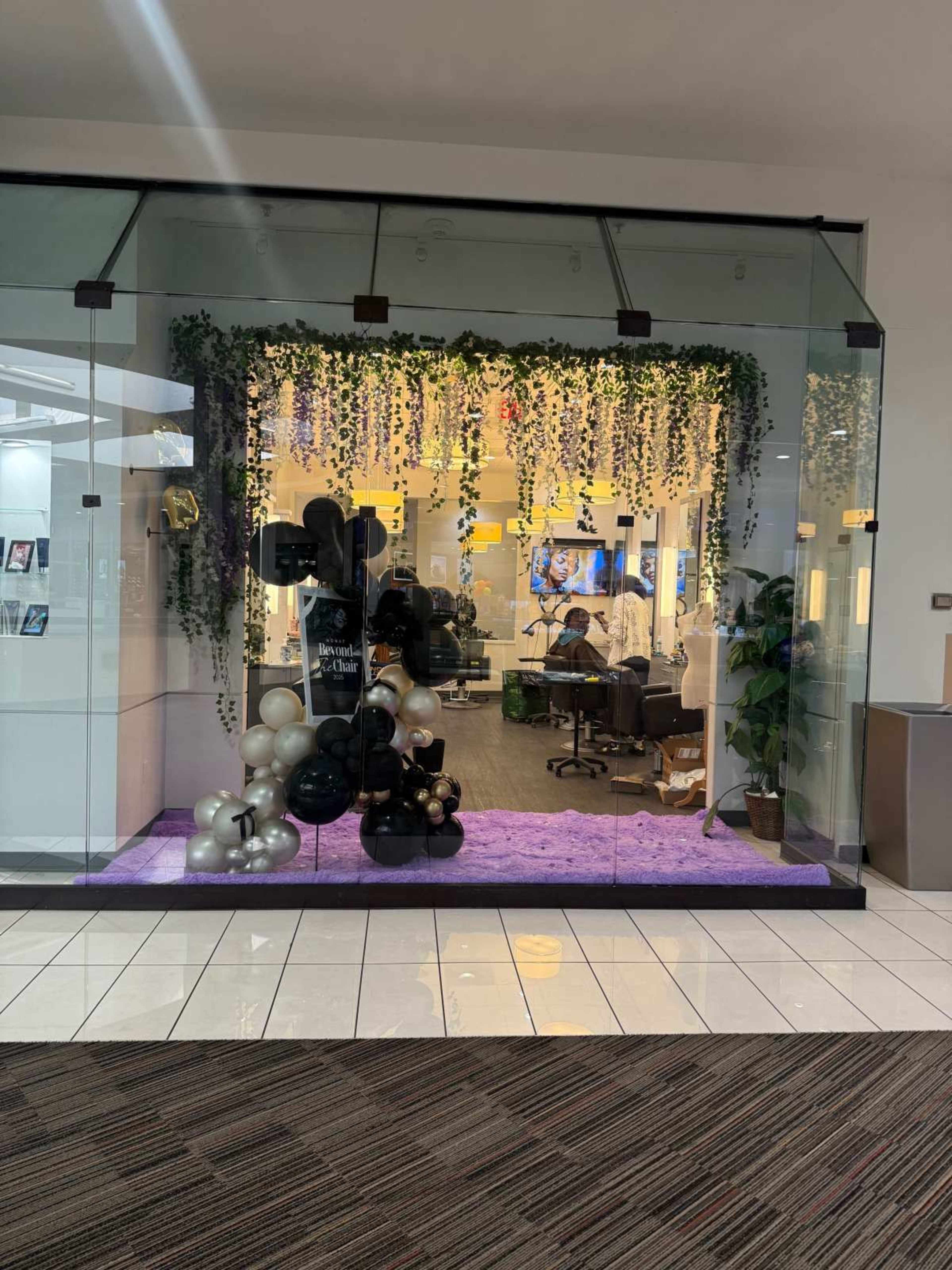 The image shows a glass storefront decorated with black and white balloons, hanging greenery, and purple carpet, with a person working inside.