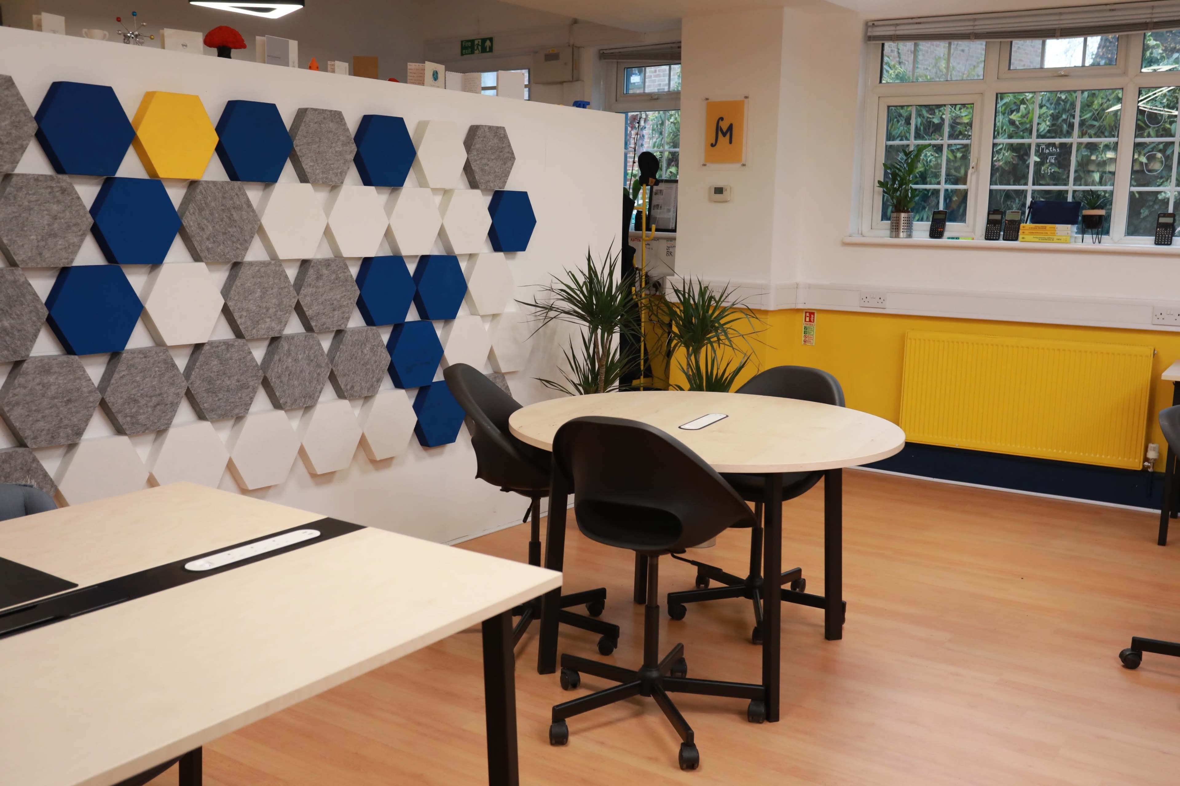 The image shows a modern classroom or meeting room with hexagonal wall panels in gray and blue, and several round tables with black chairs.