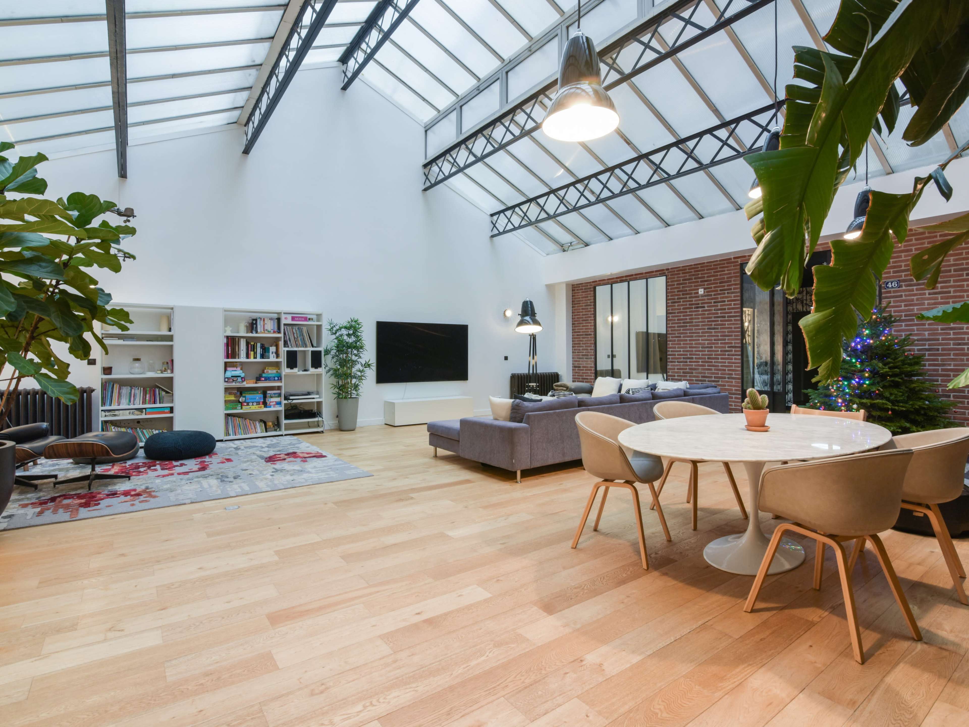 The image shows a modern, open-concept living area featuring a dining table, a sectional sofa, a large bookshelf, and floor-to-ceiling windows.