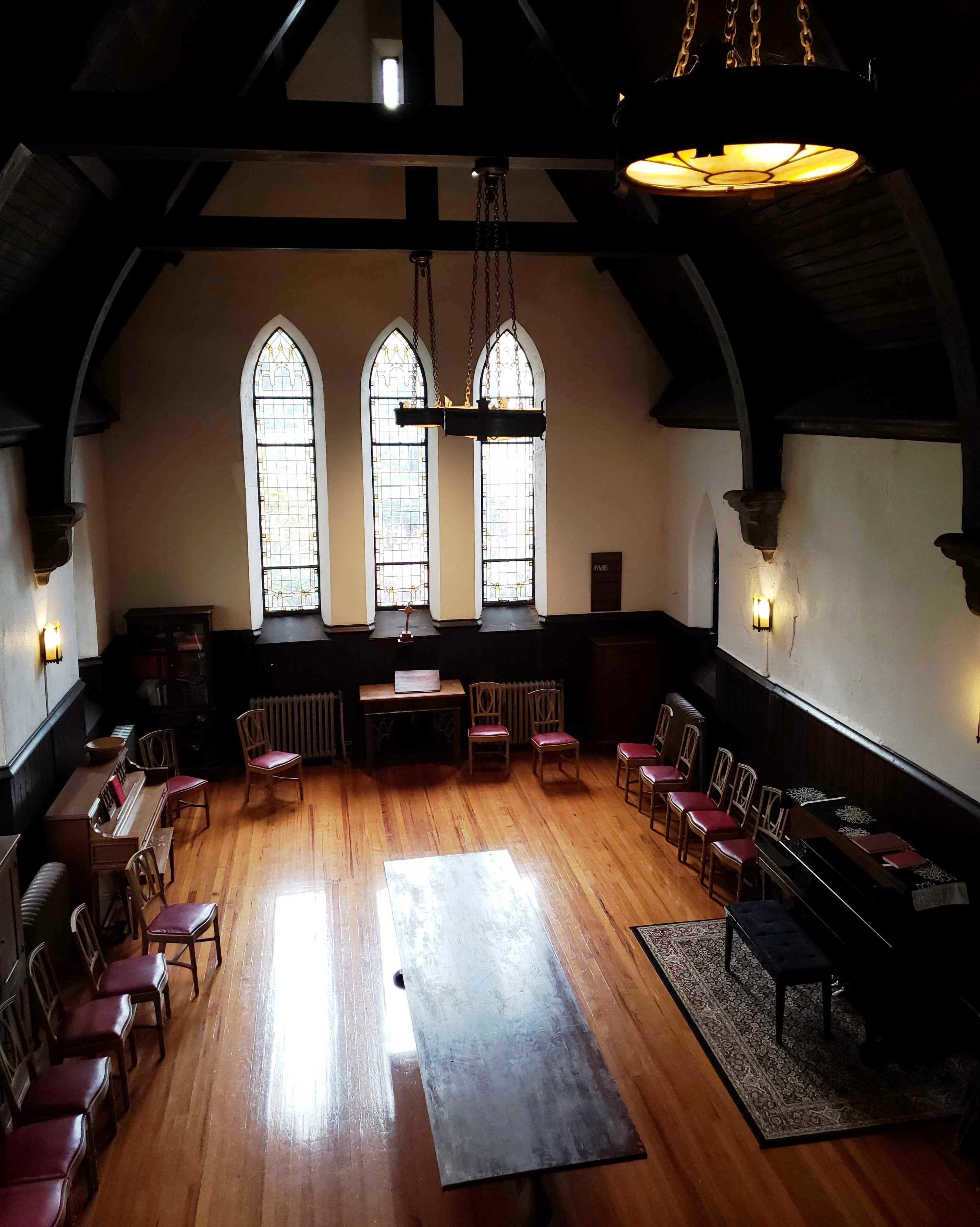 An interior room with a long wooden table surrounded by chairs, tall stained glass windows, and wooden beams in the ceiling.