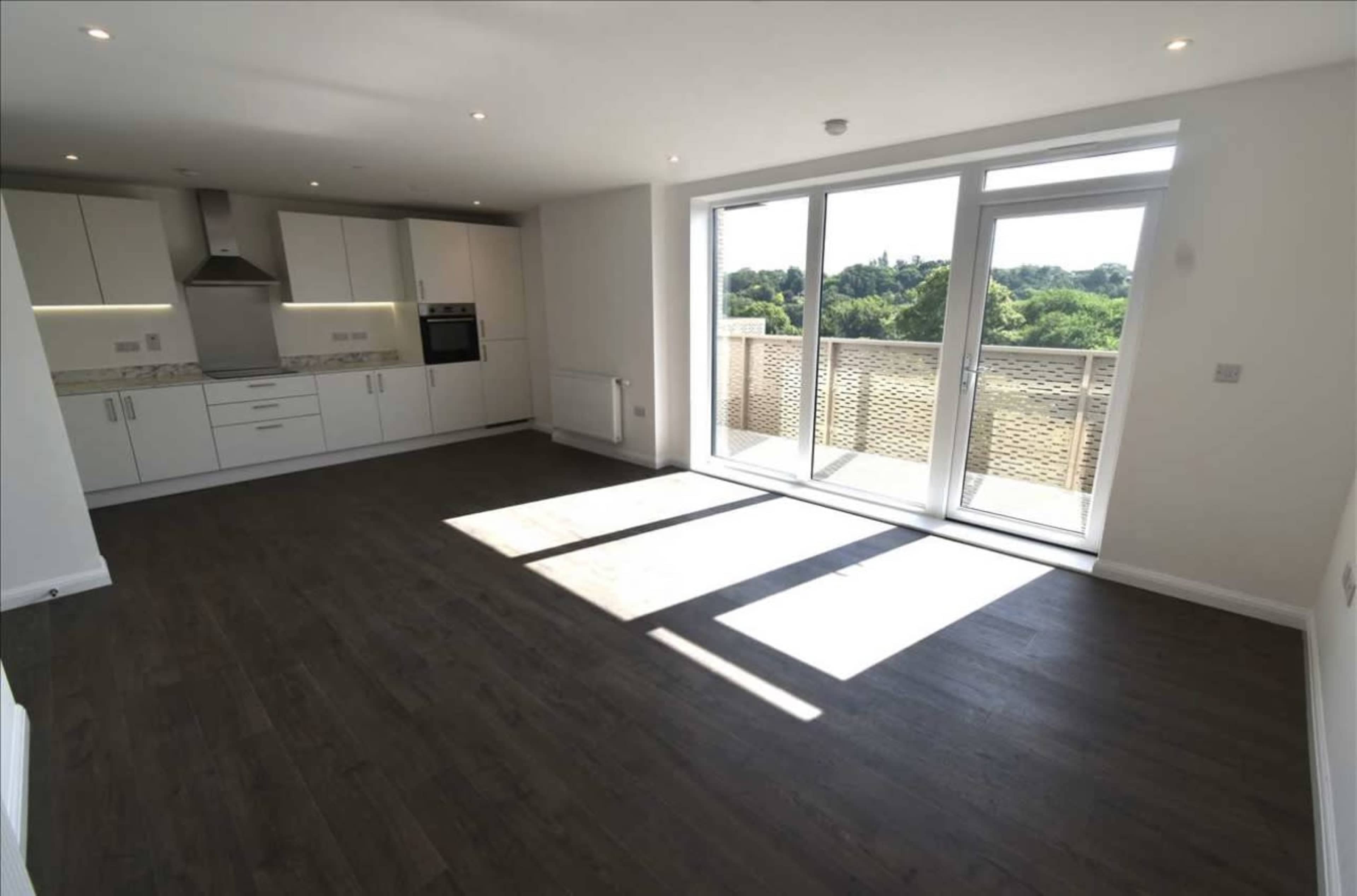 The image shows an empty room with wooden flooring, a kitchen area along one wall, and large glass doors opening to a balcony with a view of greenery outside.
