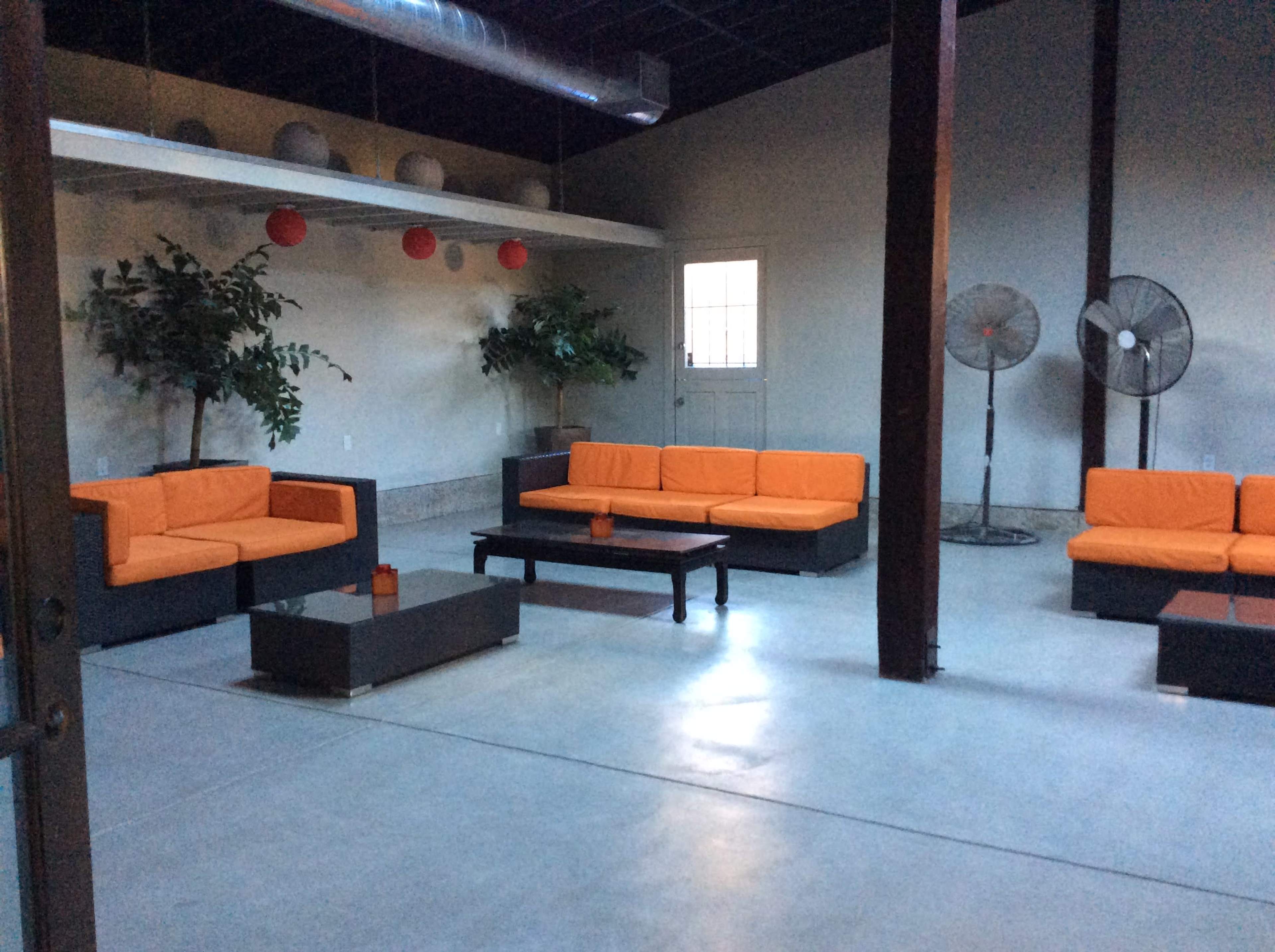 The image shows a spacious indoor area with orange sofas, a coffee table, and two large fans near the walls, under a ceiling with hanging decorations.