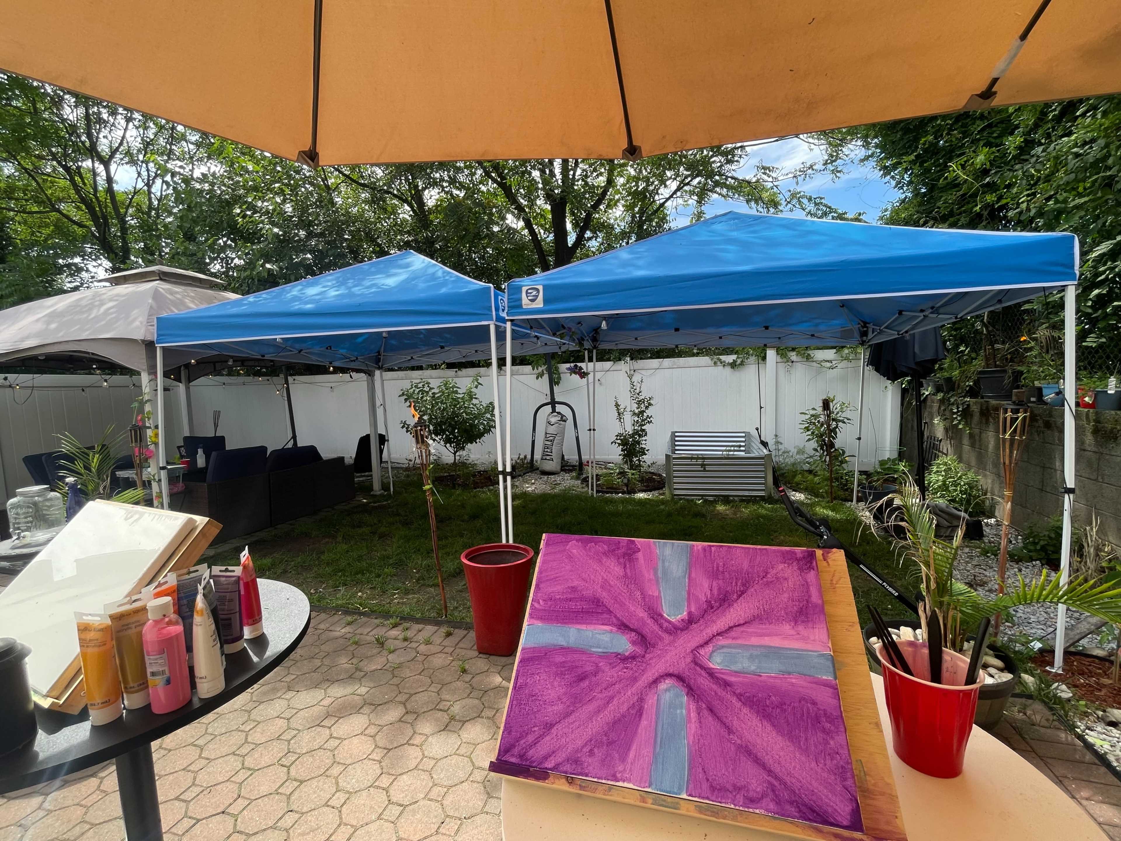 The image shows a backyard scene with two blue canopies set up, a painting on an easel, and various art supplies on a table.
