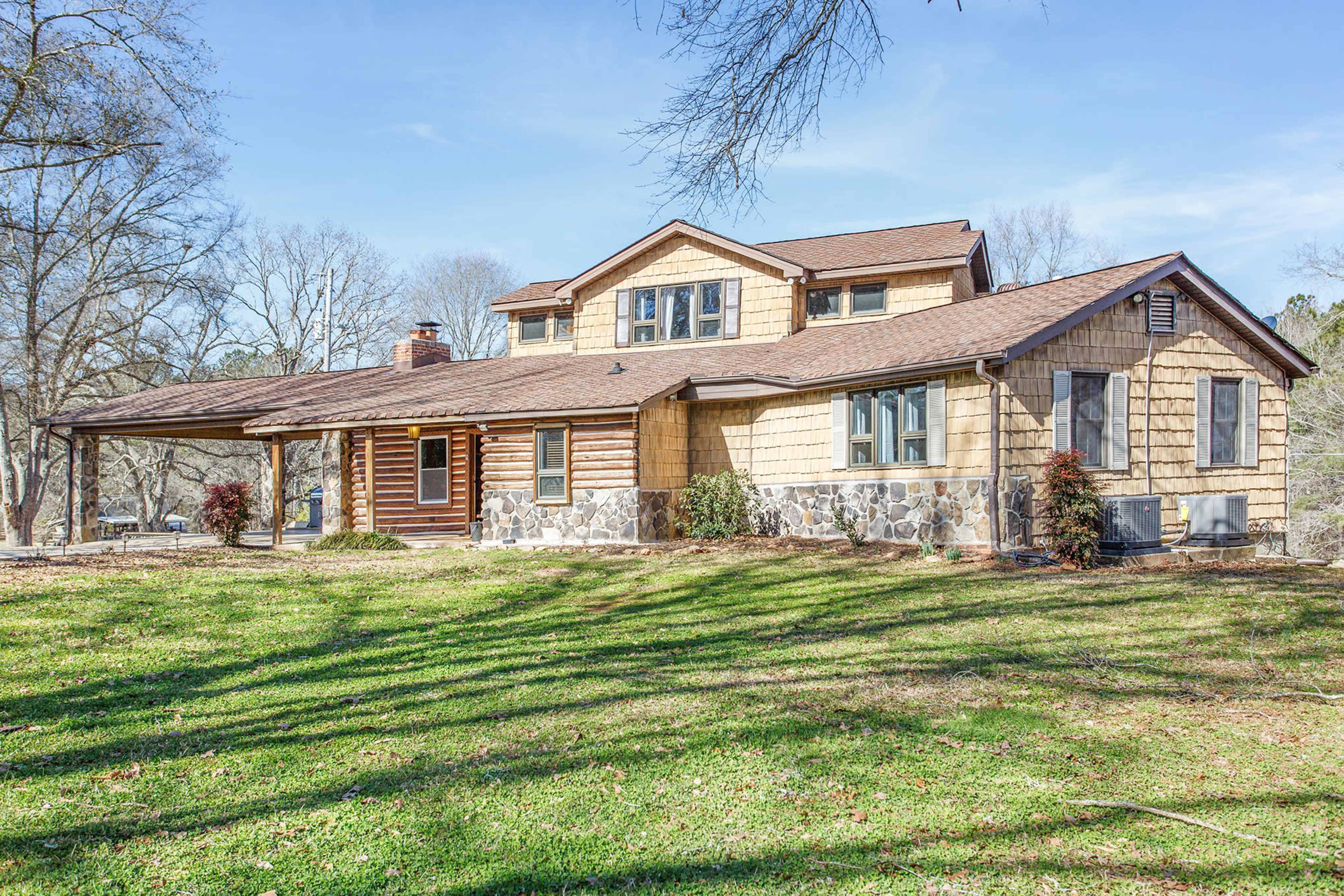 A two-story house with a combination of wooden and stone siding is situated on a grassy lawn, surrounded by trees.