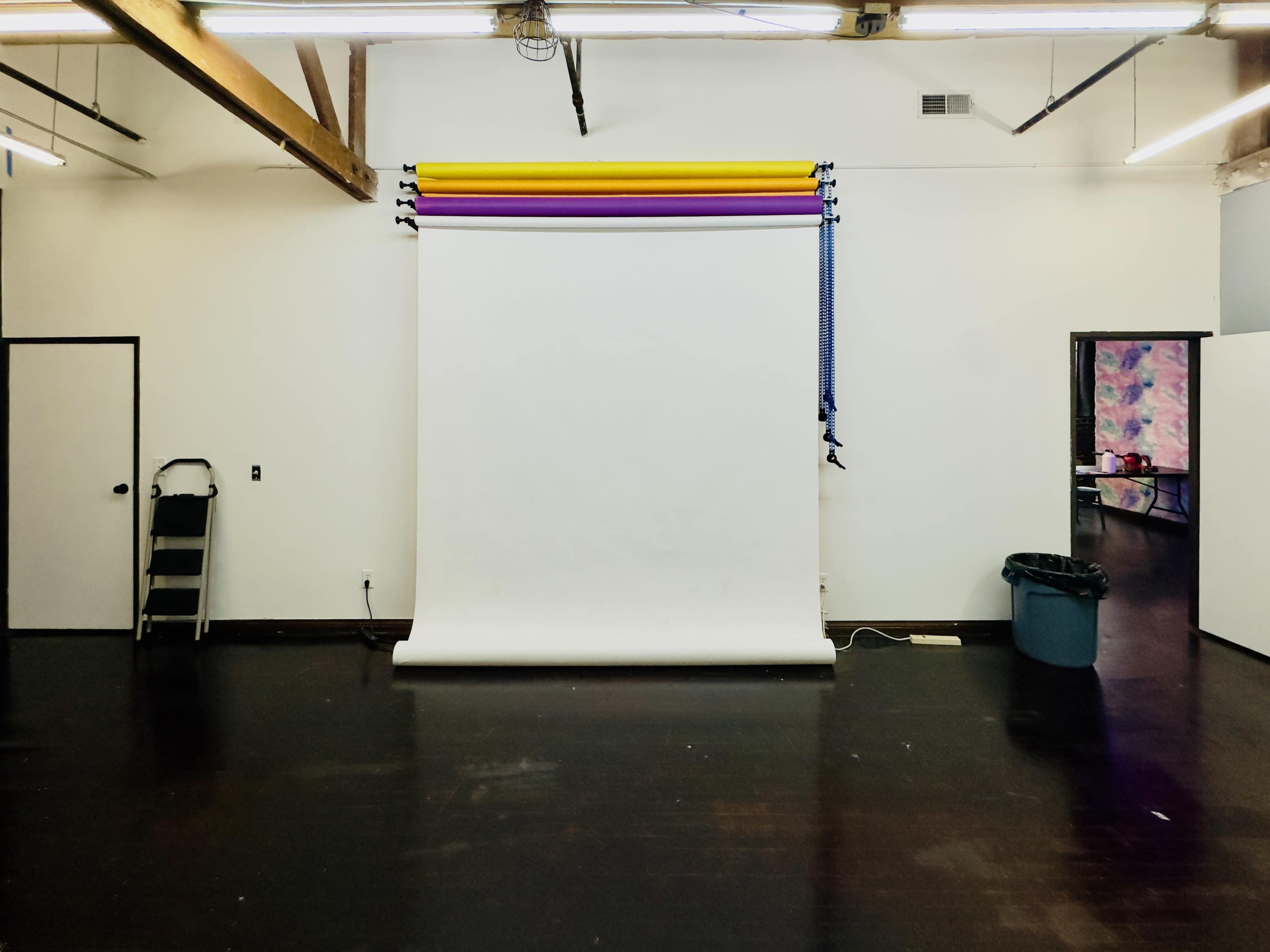 The image shows an empty studio space with a white backdrop, colorful backdrop paper, and a folding cart next to a trash bin.