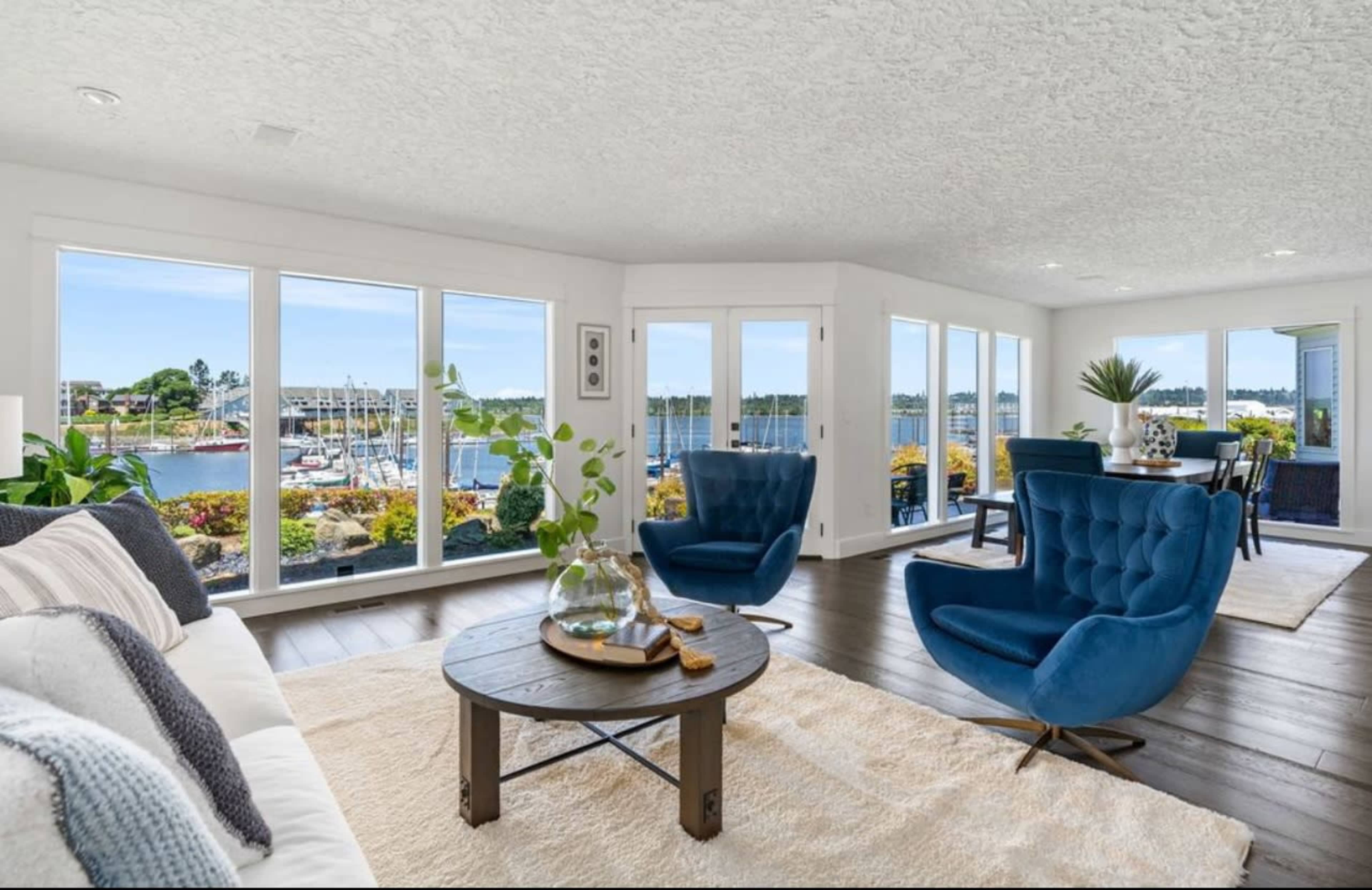 The image shows a modern living room with large windows offering a view of a marina filled with boats, featuring blue chairs, a round table, and a light-colored rug.