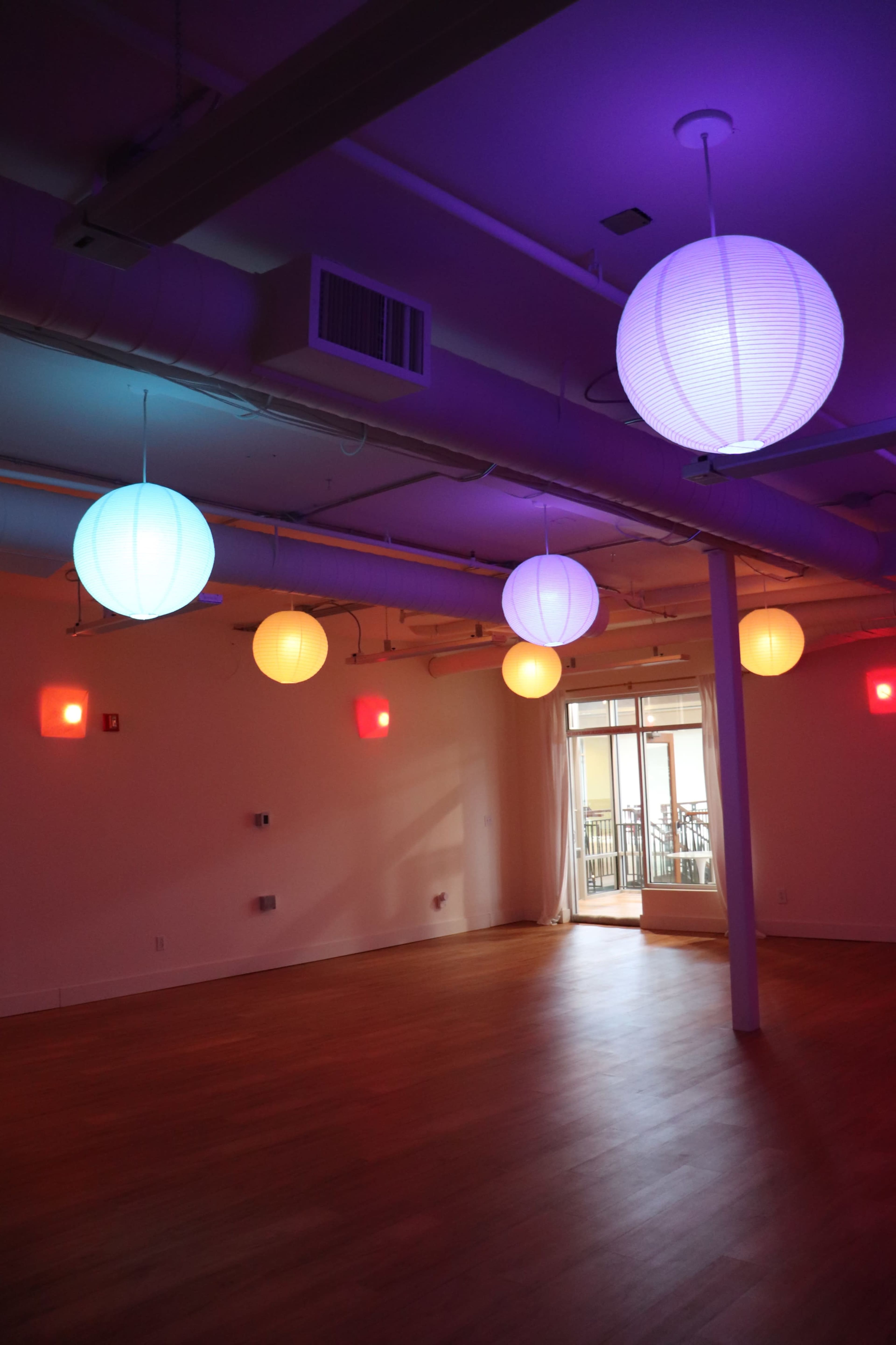 The image shows a spacious room with wooden flooring, illuminated by multiple hanging paper lanterns in various colors and two red wall lights.