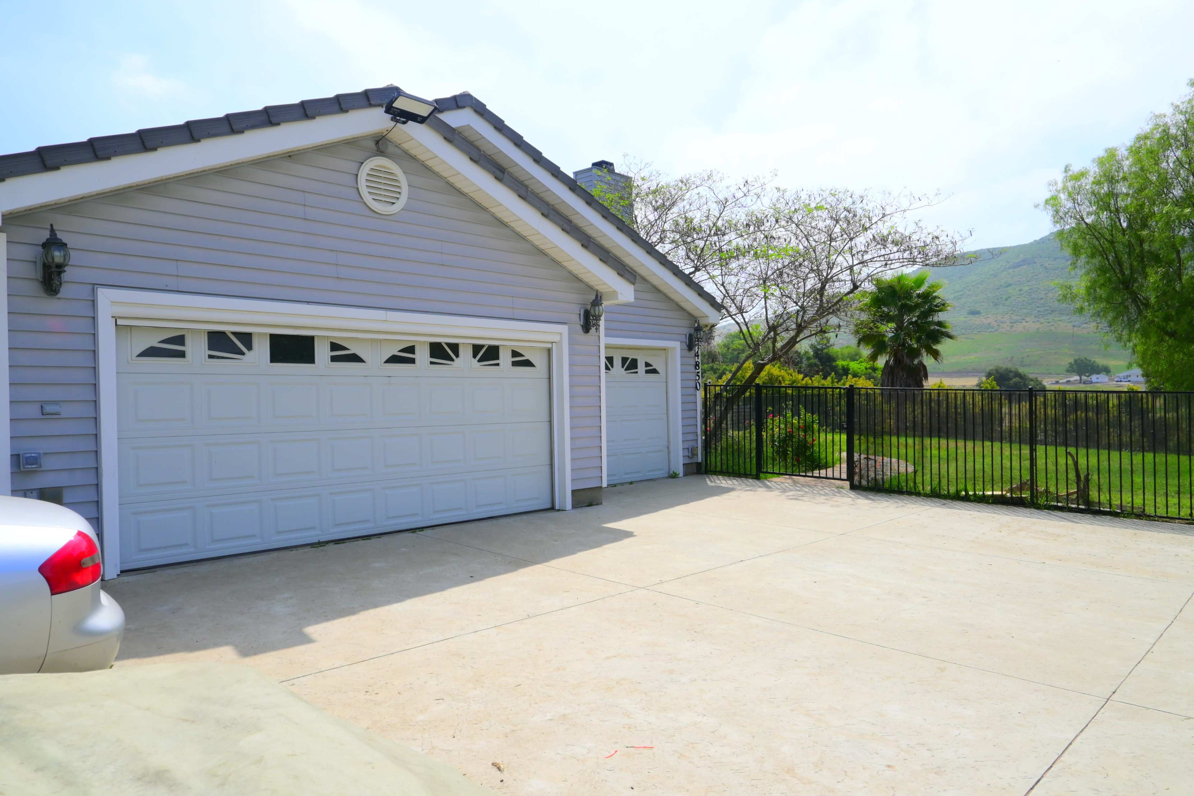 A gray house with a two-car garage faces a concrete driveway and is surrounded by a metal fence and greenery.