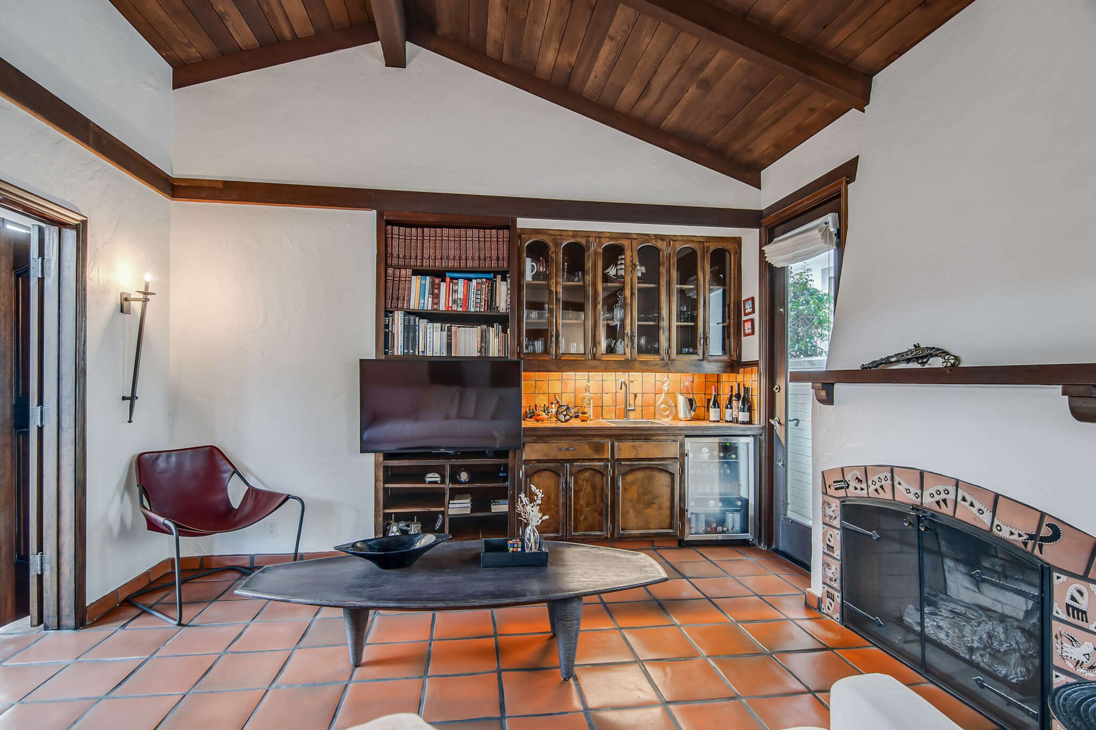 A cozy living room with a wooden ceiling, tiled floor, built-in bookshelves, a television, and a fireplace.