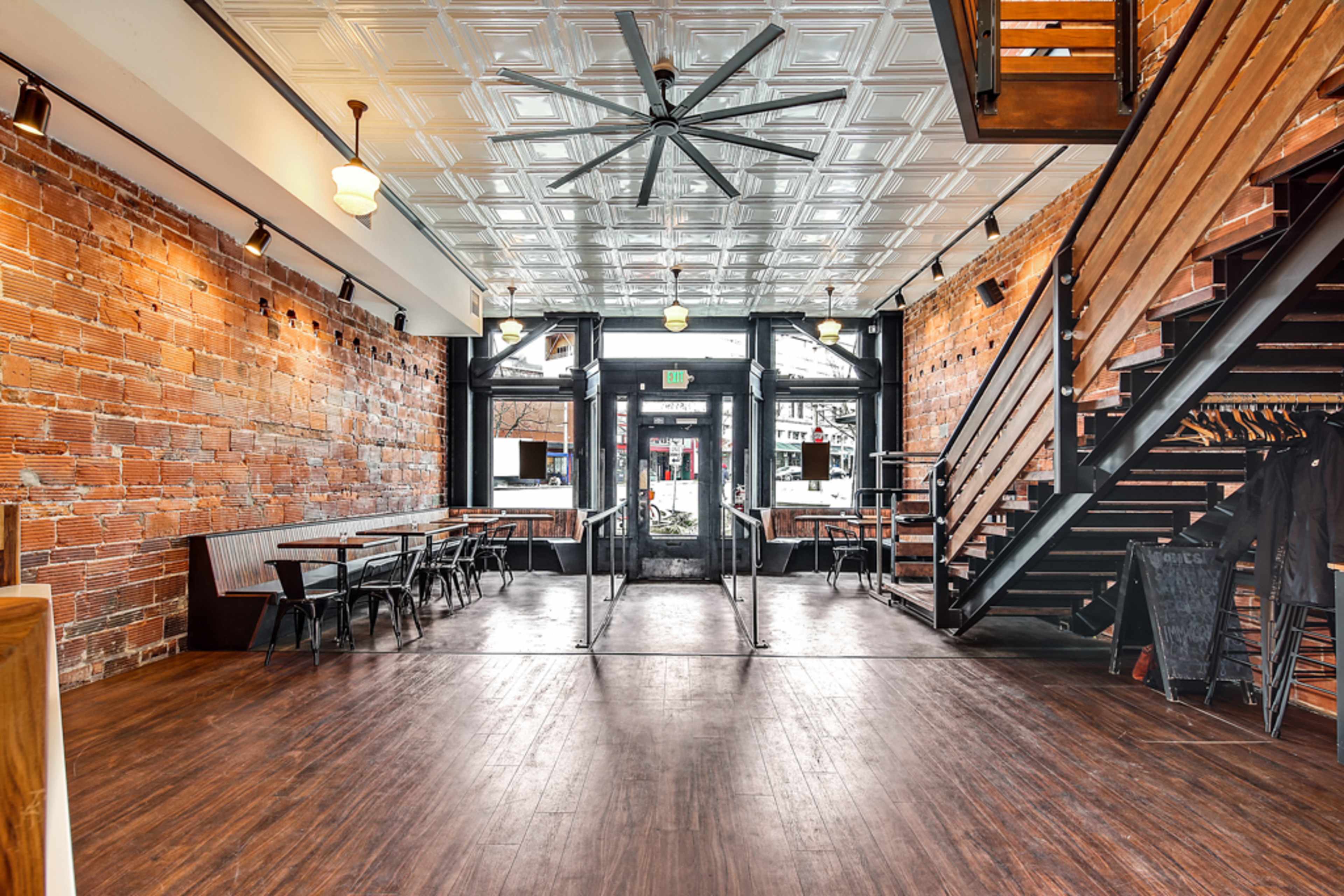 A spacious interior of a restaurant featuring exposed brick walls, wooden floors, and a large ceiling fan, with tables and a staircase visible.