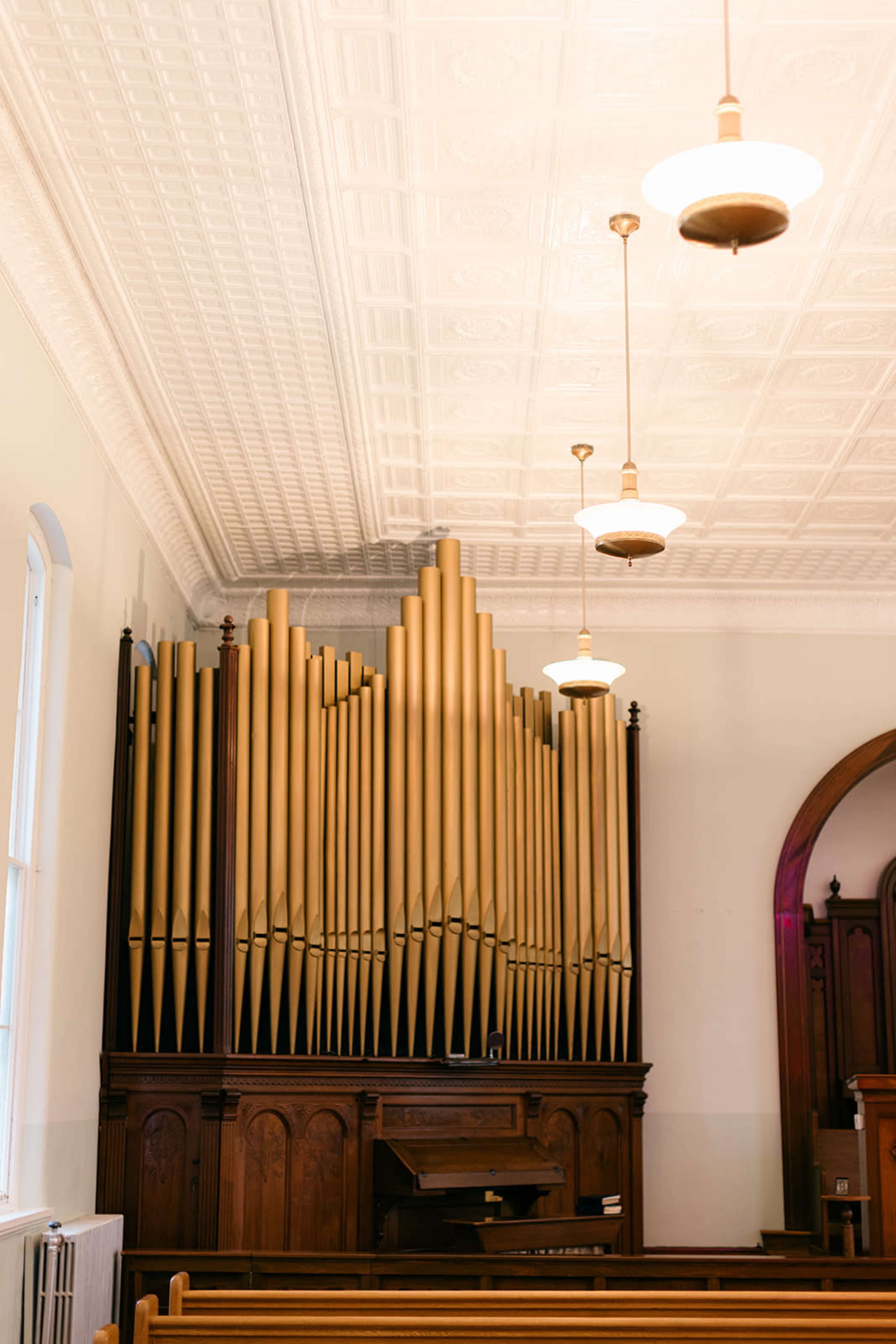 The image shows a church interior featuring a large pipe organ against a wall, with wooden pews and pendant lighting.