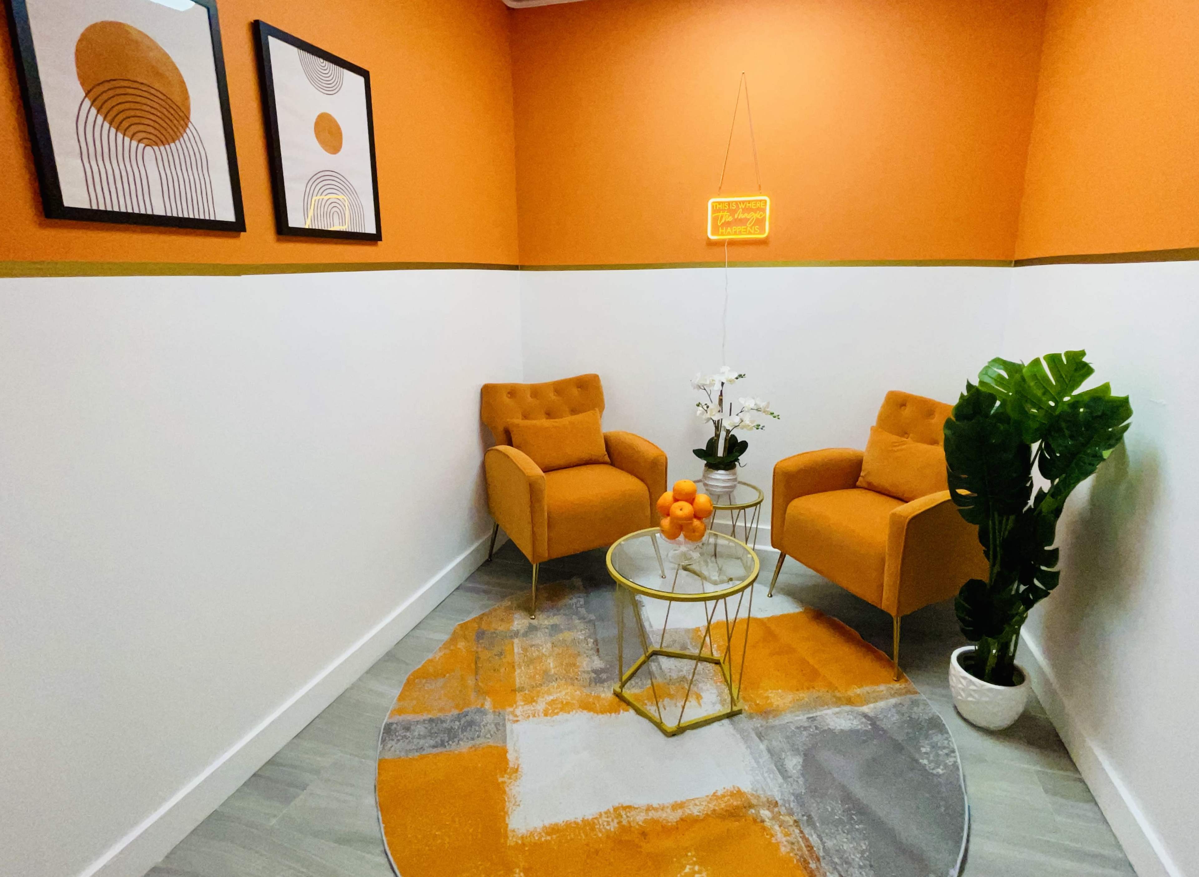 A small, modern room with orange walls, two orange armchairs, a round coffee table, and decorative plant elements.