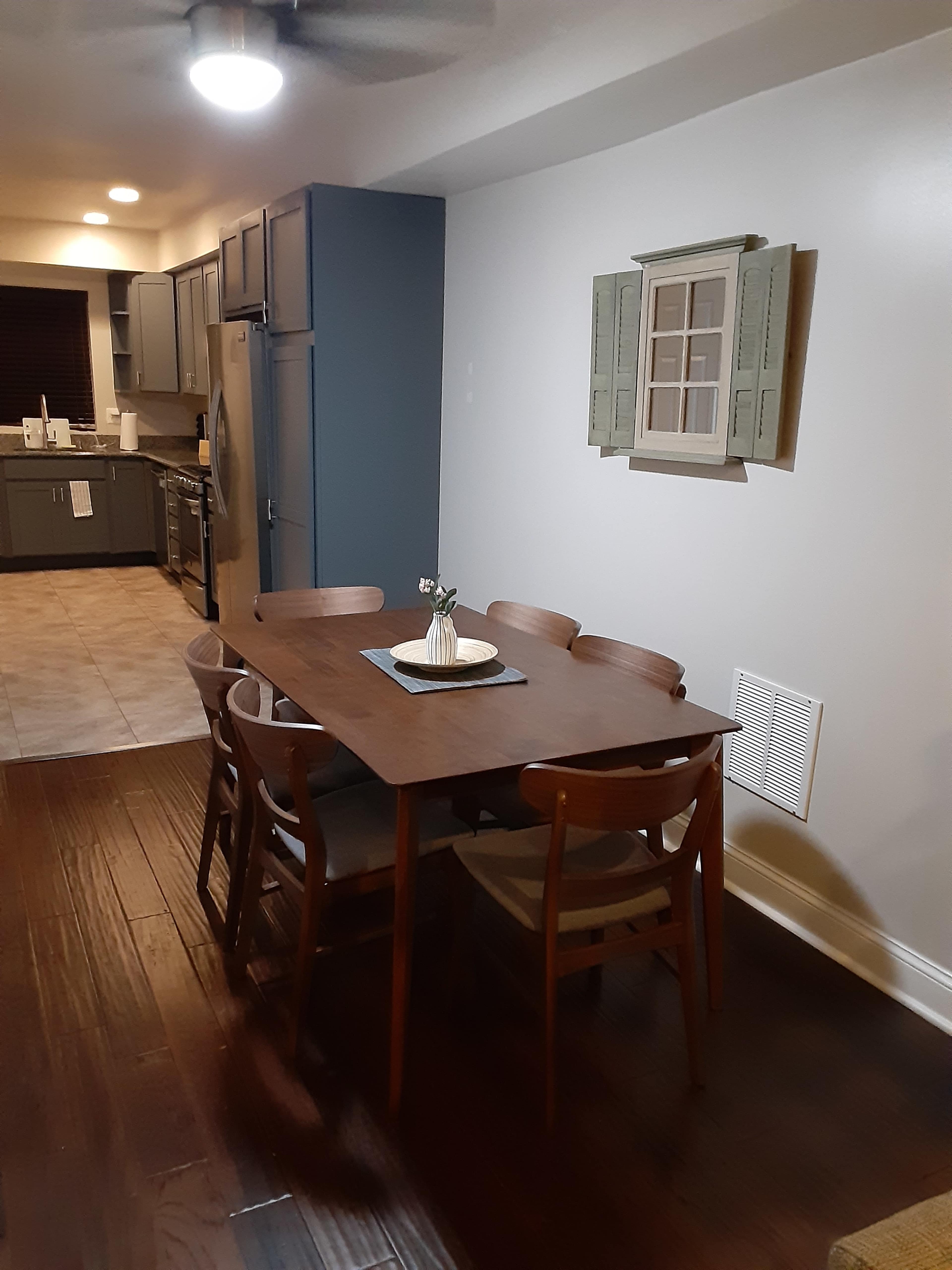 A wooden dining table with six chairs is positioned in a well-lit kitchen featuring gray cabinetry and a decorative window.