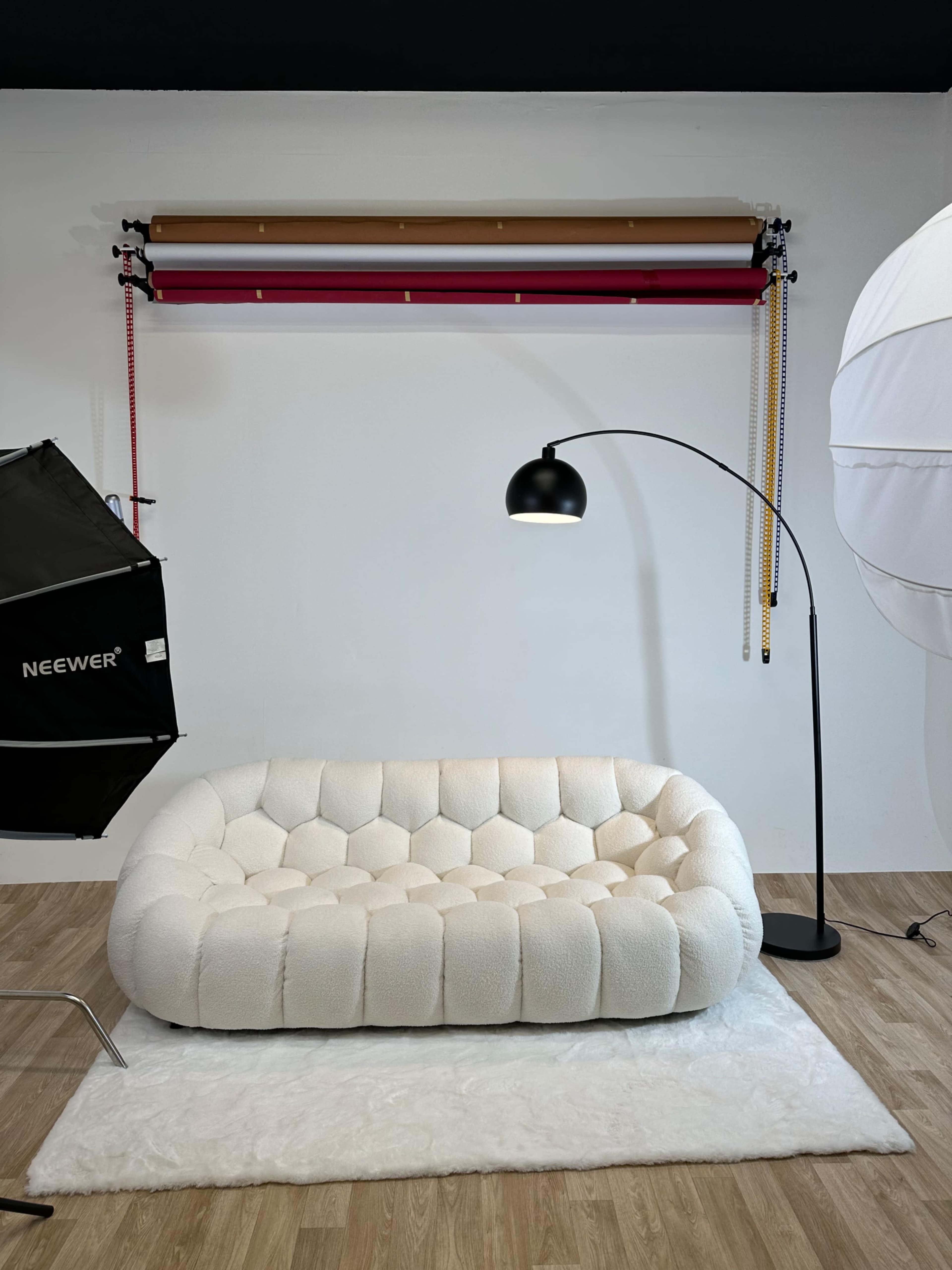 The image features a modern, upholstered sofa on a white rug, positioned in front of a plain wall, with a backdrop support system and a studio light setup in the surrounding area.
