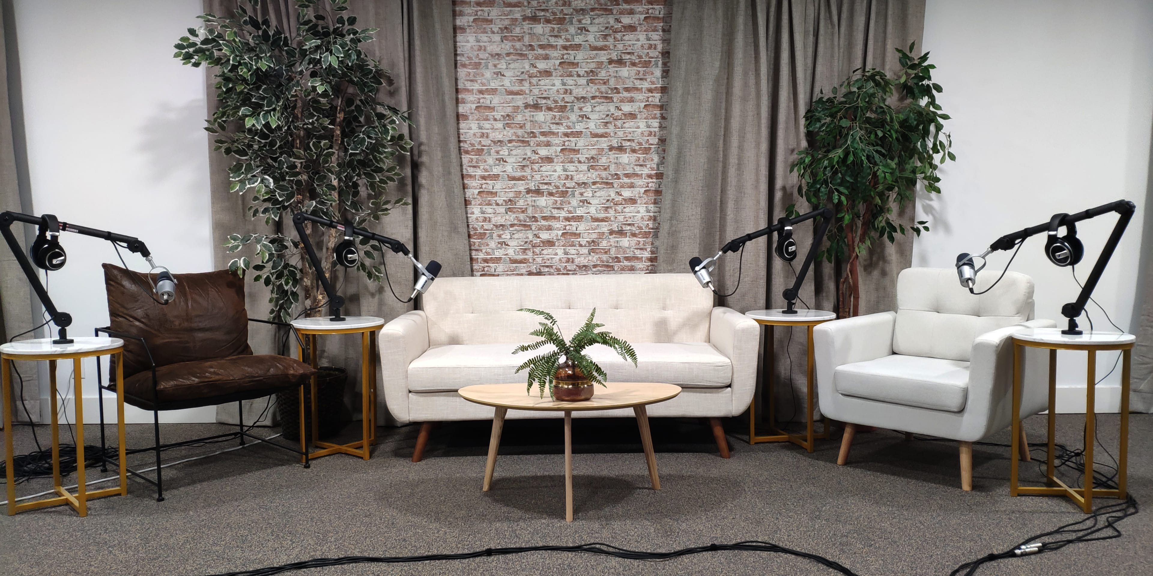 A small, professionally arranged interview or talk show set featuring a white sofa, two armchairs, potted plants, and microphone stands on either side.