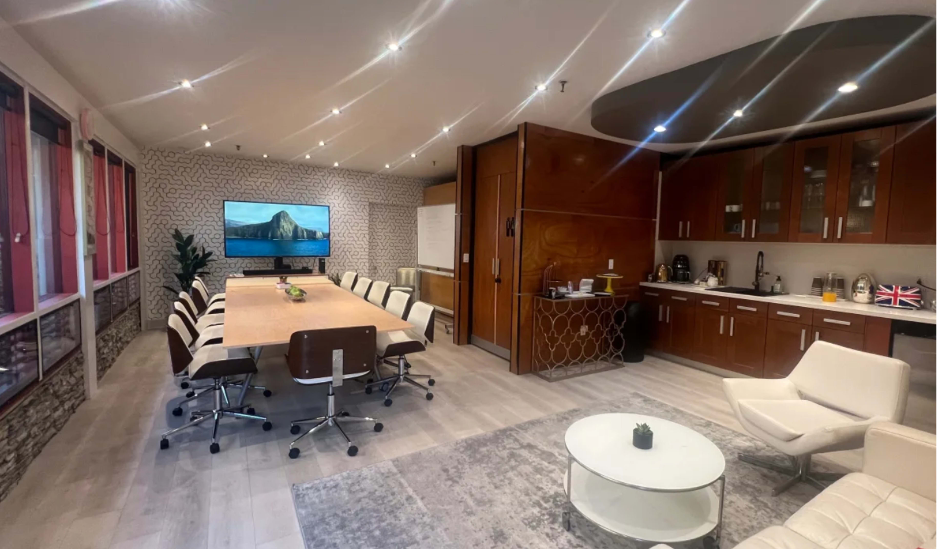 A modern conference room with a large table surrounded by ergonomic chairs, a wall-mounted TV, and a kitchenette area.