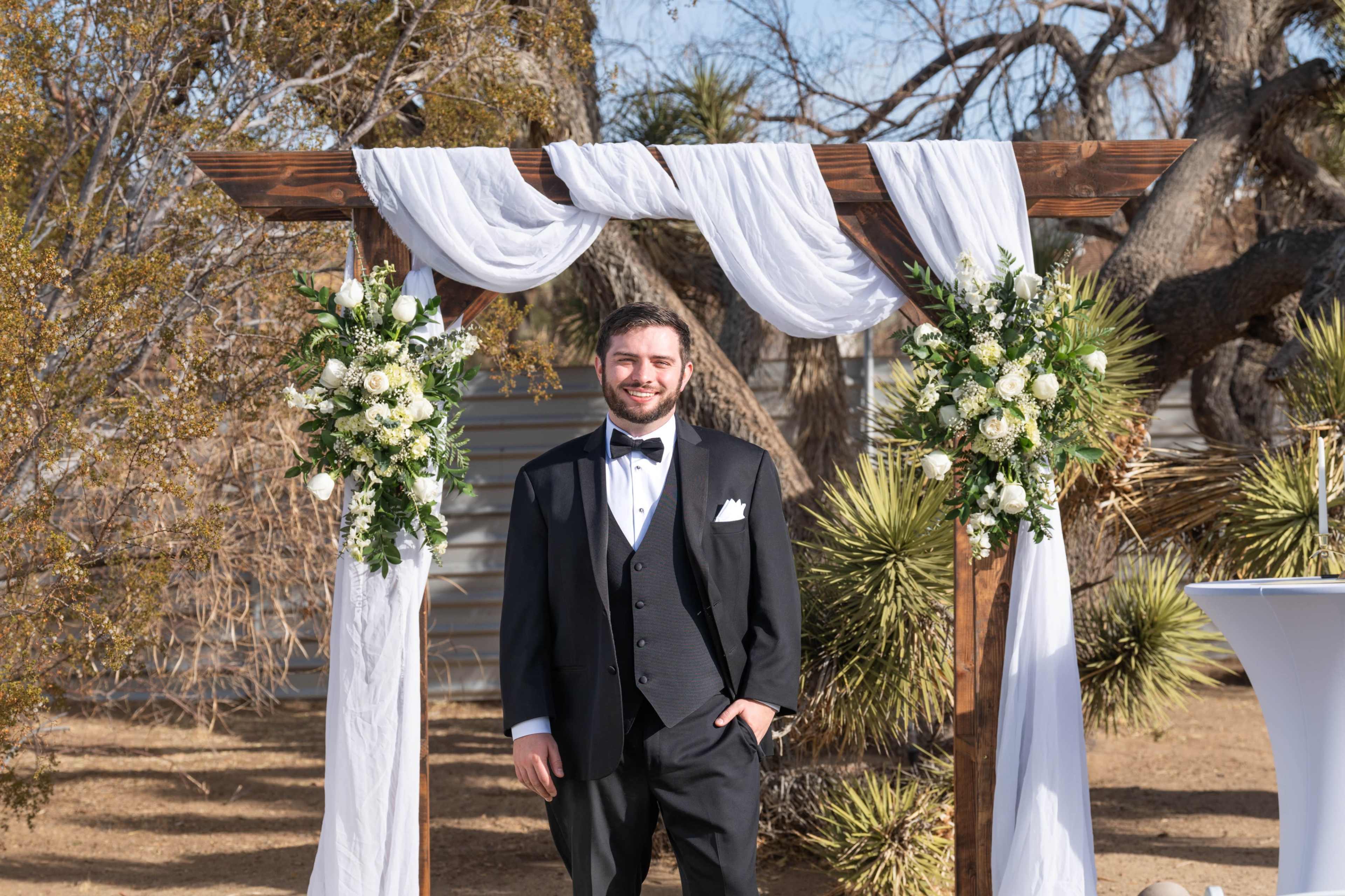 A man in a tuxedo stands beneath a decorative wedding arch adorned with white drapery and flowers in a desert setting.
