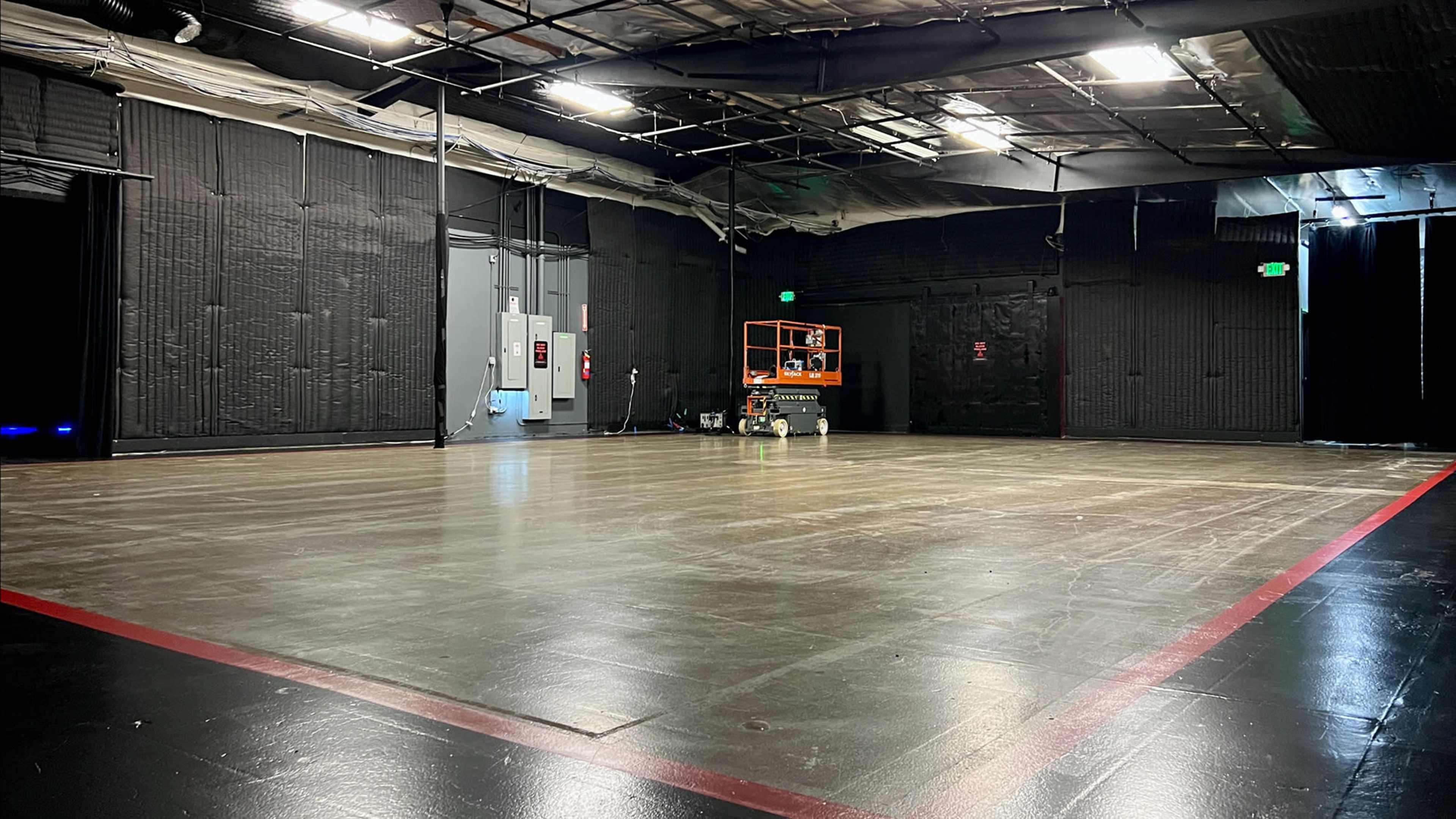 The image shows a spacious, empty film studio with a smooth floor, overhead lights, and equipment in one corner.