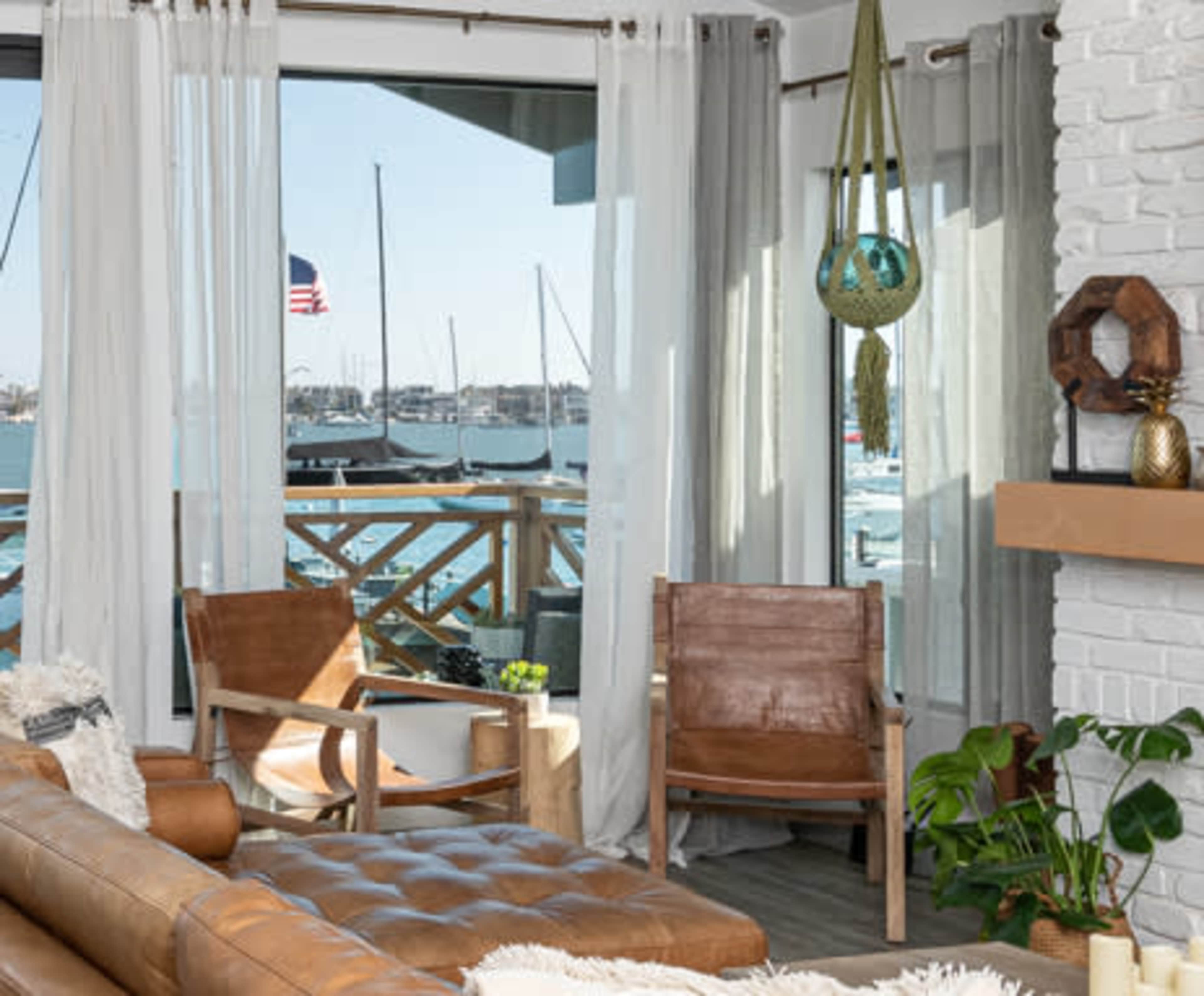 The scene features a cozy living room with large windows revealing a harbor filled with sailboats, framed by sheer curtains and two leather chairs.