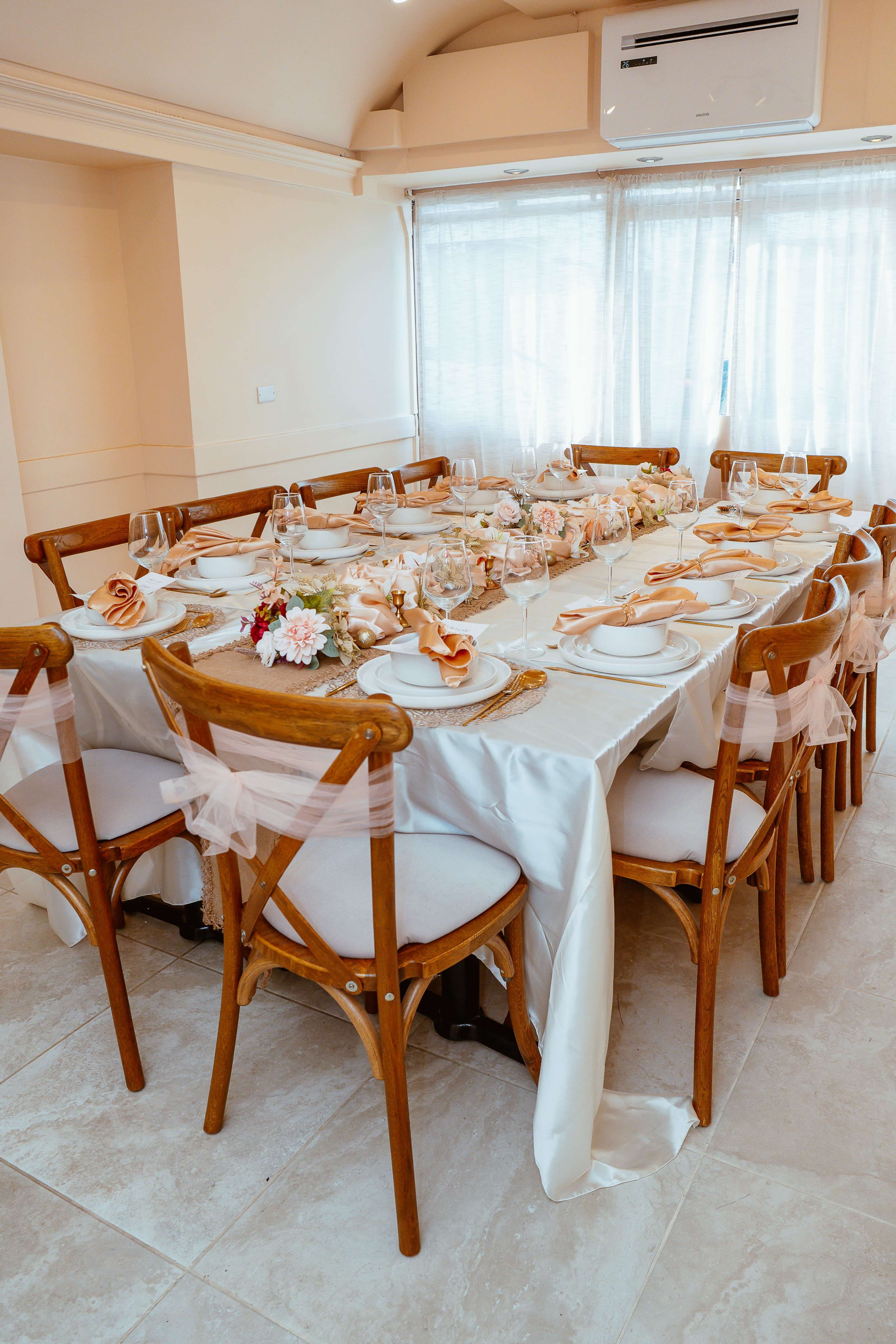 A long dining table is elegantly set with place settings and decorative floral arrangements under a well-lit window.