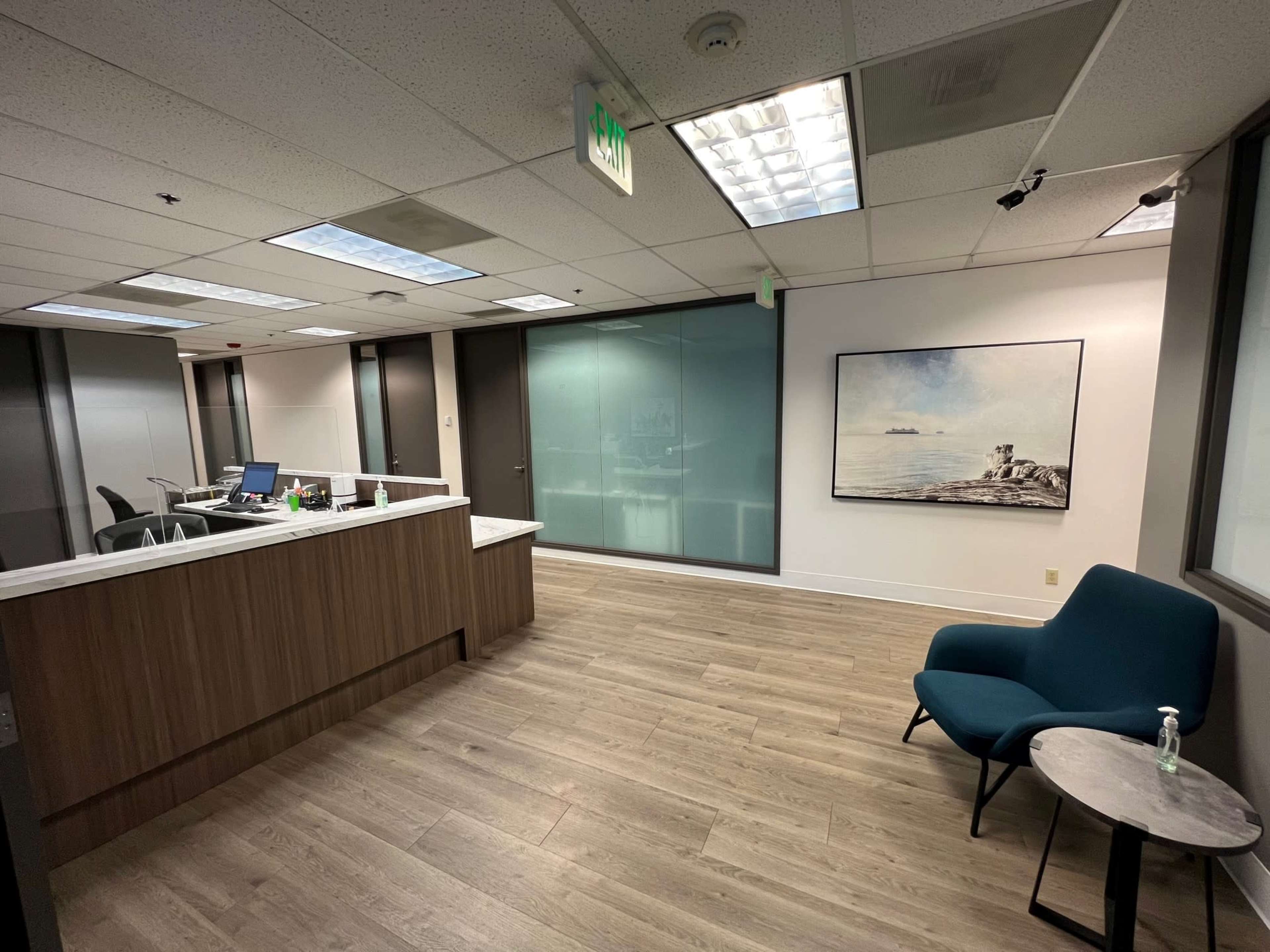 A modern office space features a reception area with a wooden counter, a blue chair, and a large artwork on the wall, along with a glass-walled meeting room.