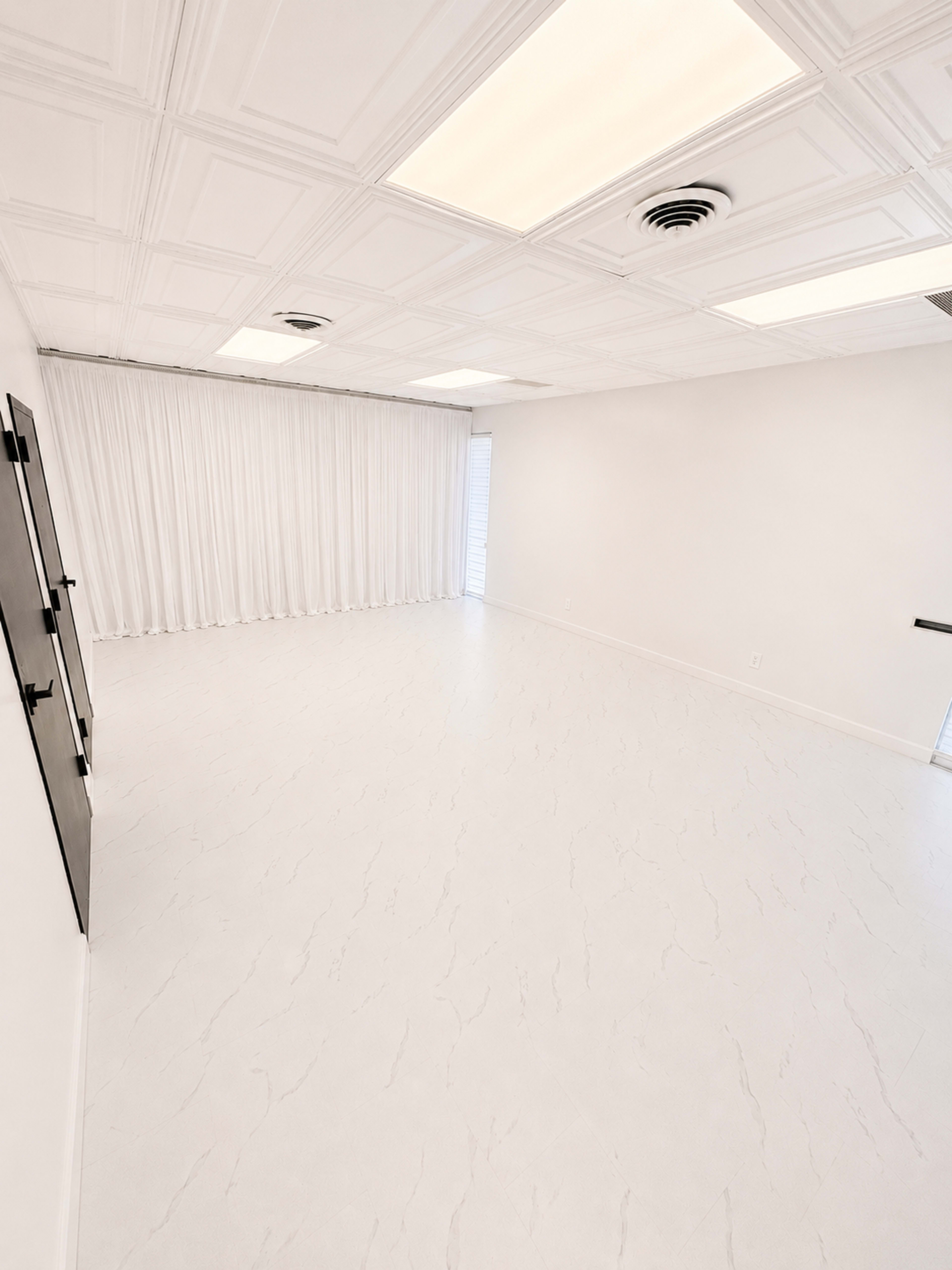 The image depicts a spacious, minimalist room with a white textured floor and bright ceiling lights, featuring a large window covered by sheer curtains.