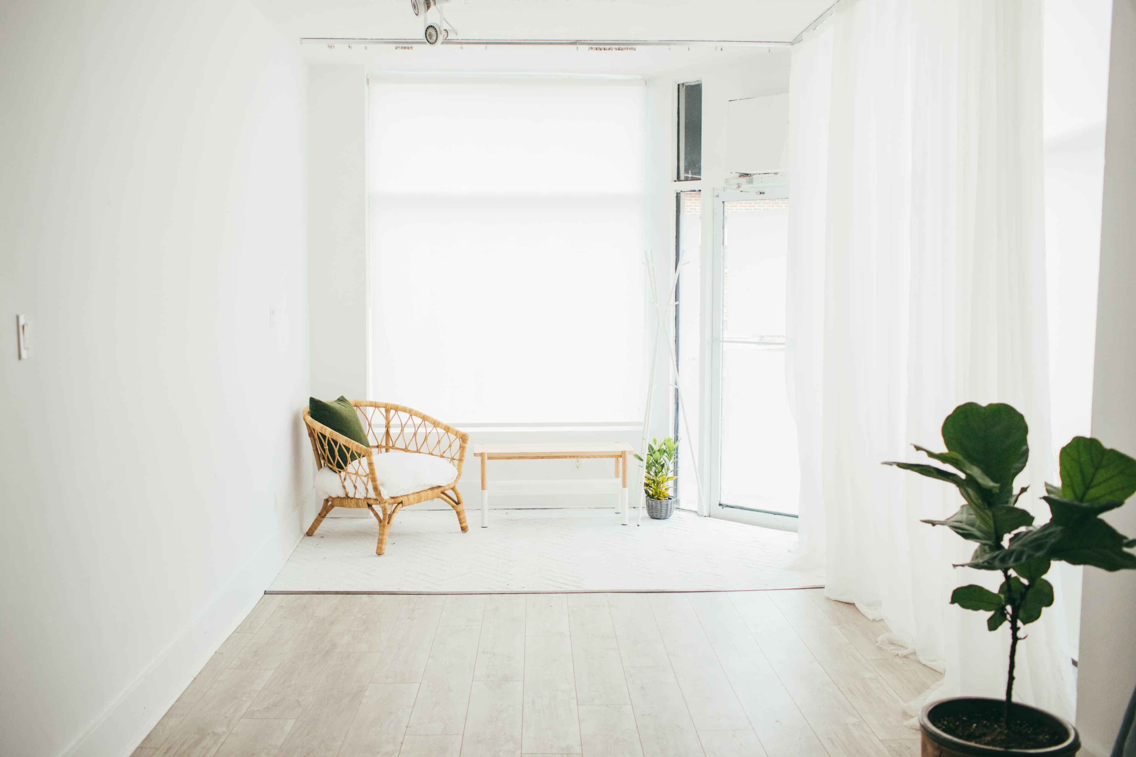 A bright, minimalistic room features a wicker chair and a small table beside a large window with sheer curtains, complemented by a potted plant.