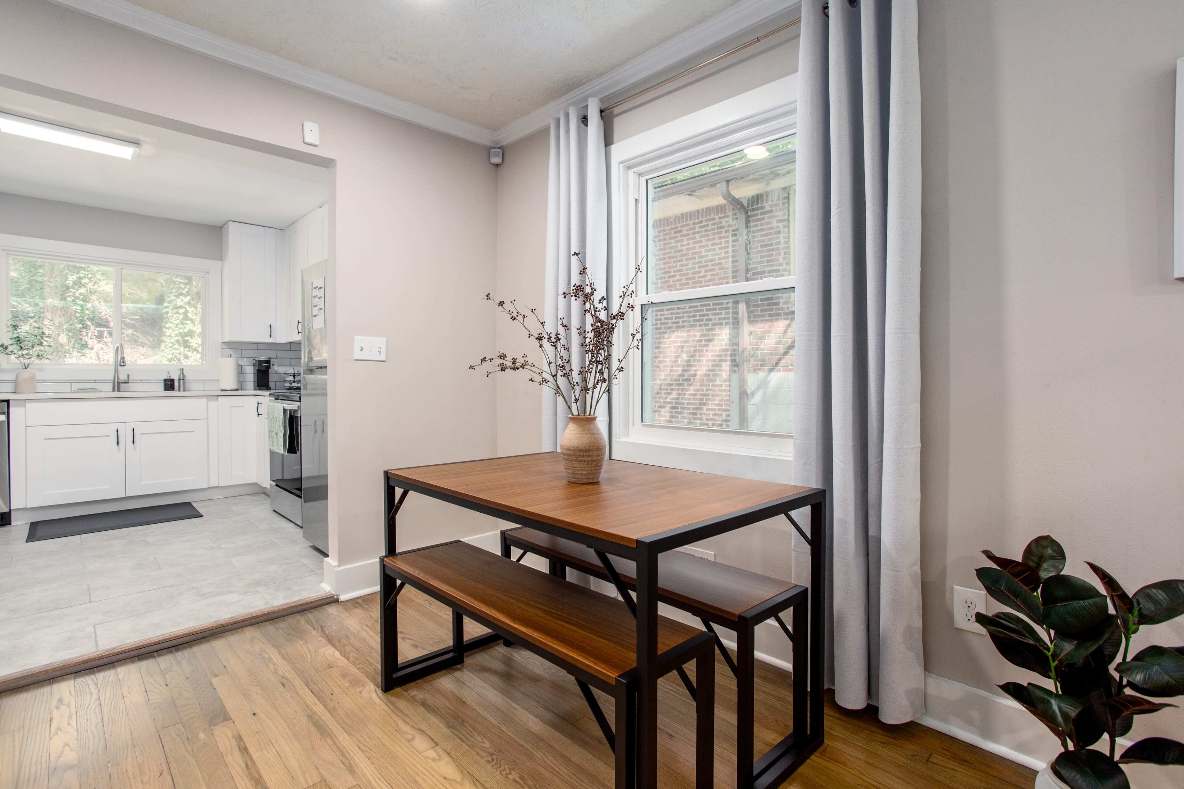 A wooden table with benches is situated in a bright dining area, adjacent to a kitchen with modern appliances and a window overlooking greenery.
