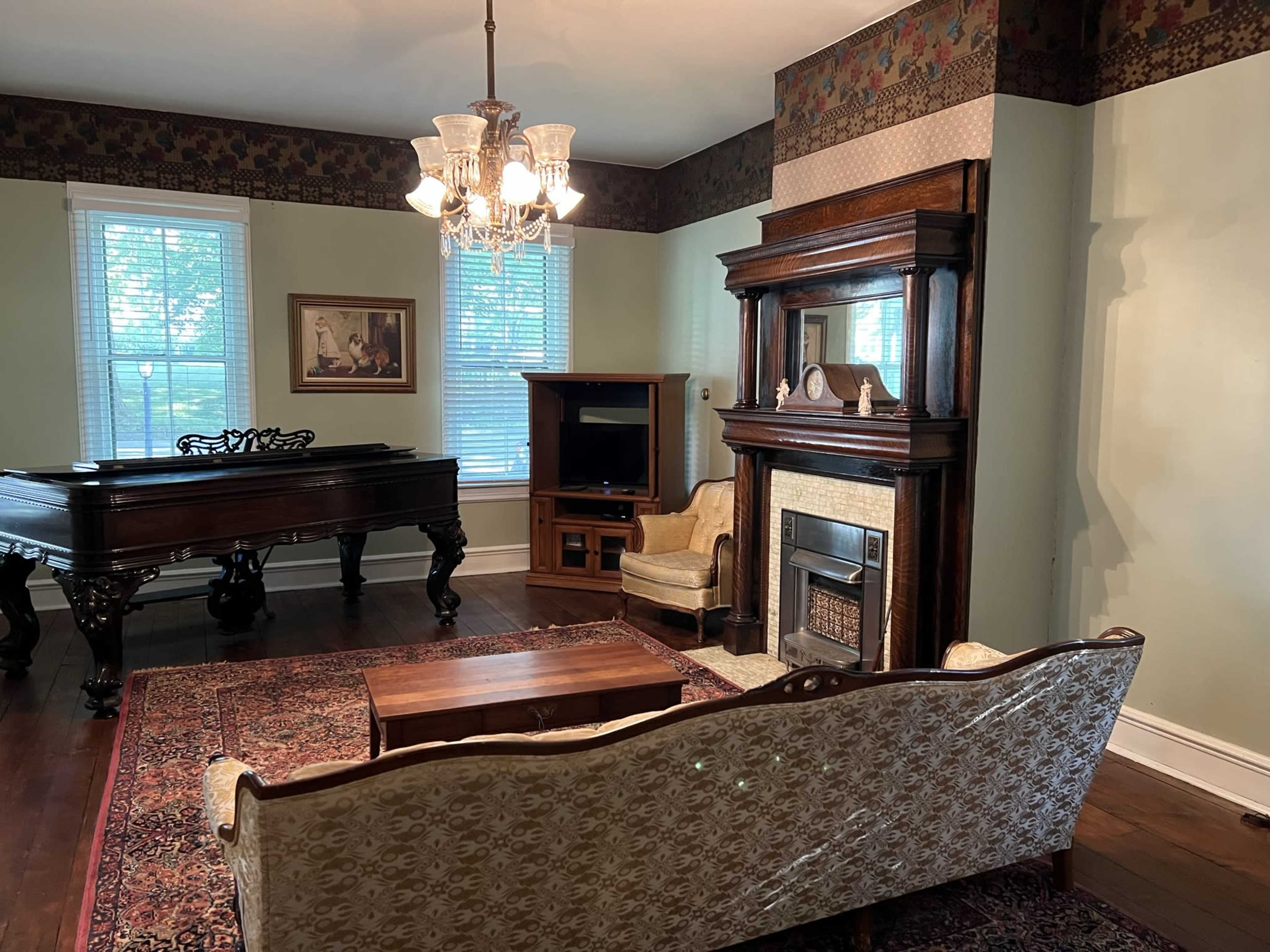 The room features a vintage-style seating area with a large ornate fireplace, a piano, and large windows letting in natural light.