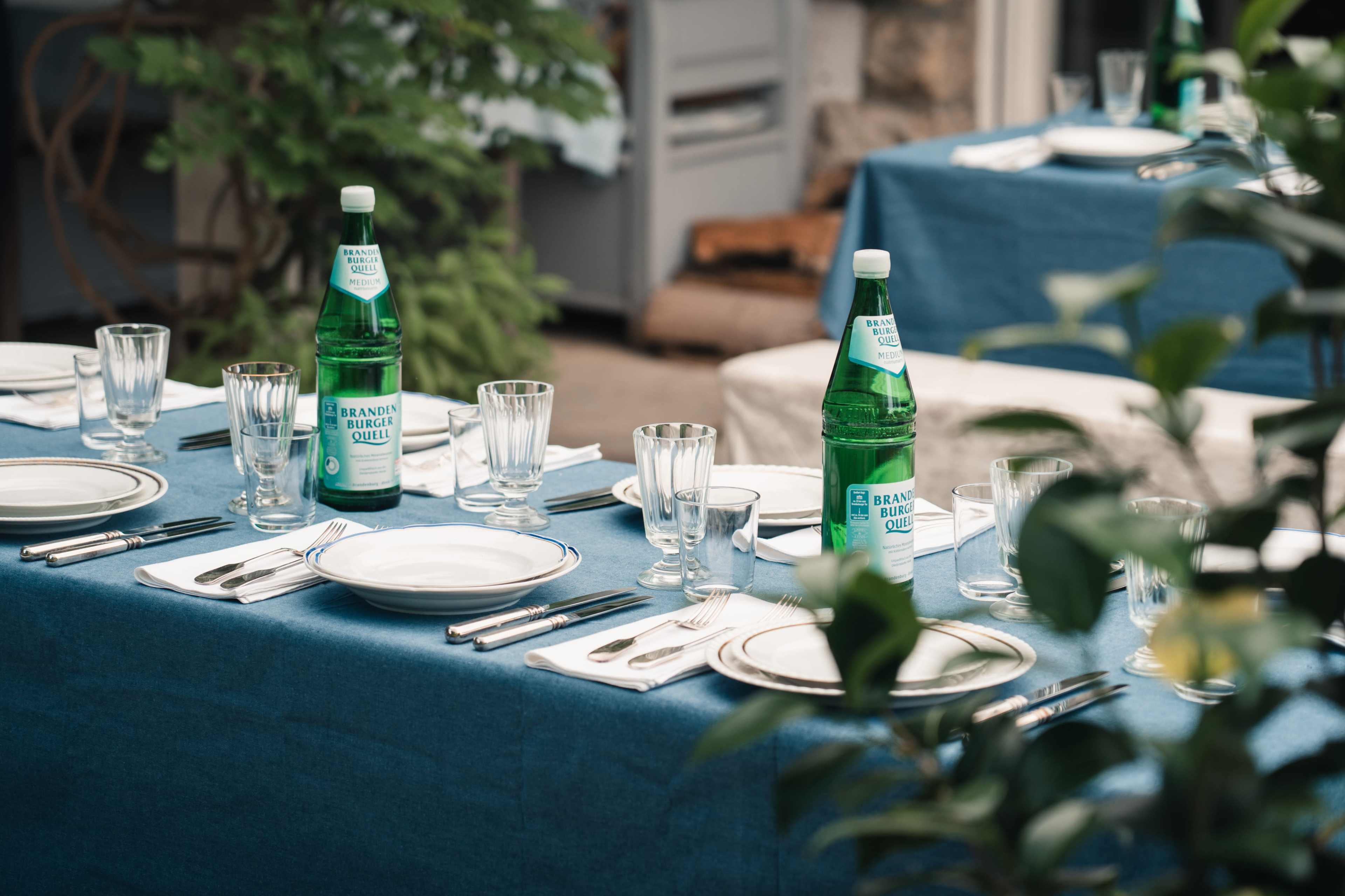 The image shows a dining setup with blue tablecloths, water bottles, glassware, and plates arranged neatly on a table in an outdoor setting.