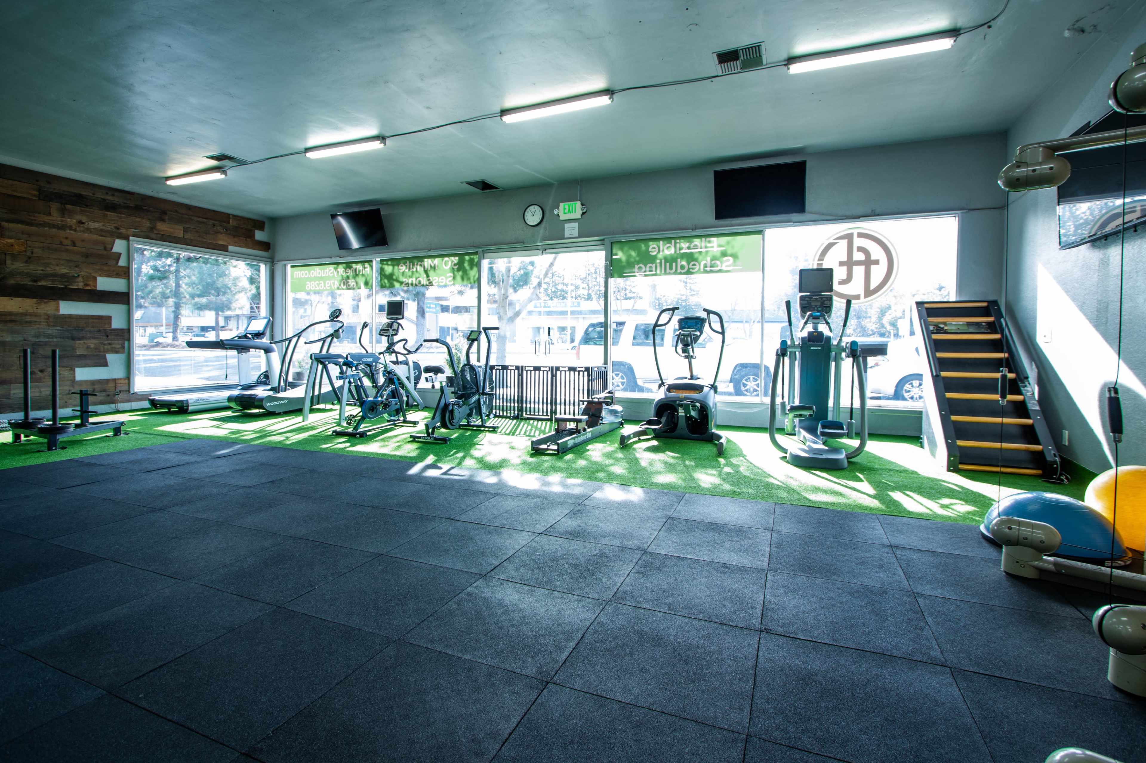 A gym interior with various exercise equipment, including cardio machines and weights, situated on rubber flooring and illuminated by natural light.