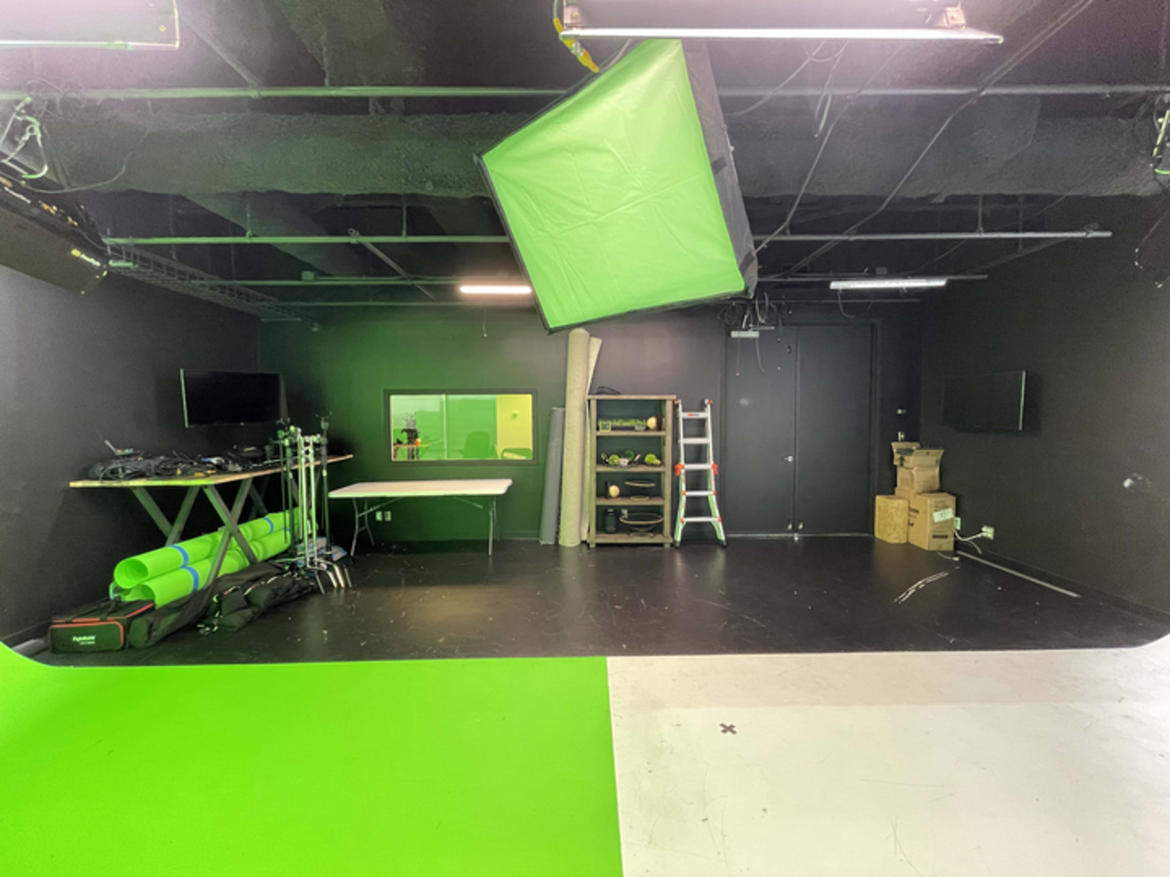 A studio space with a green screen setup, black walls, a table, some equipment on the floor, and a ladder against the wall.