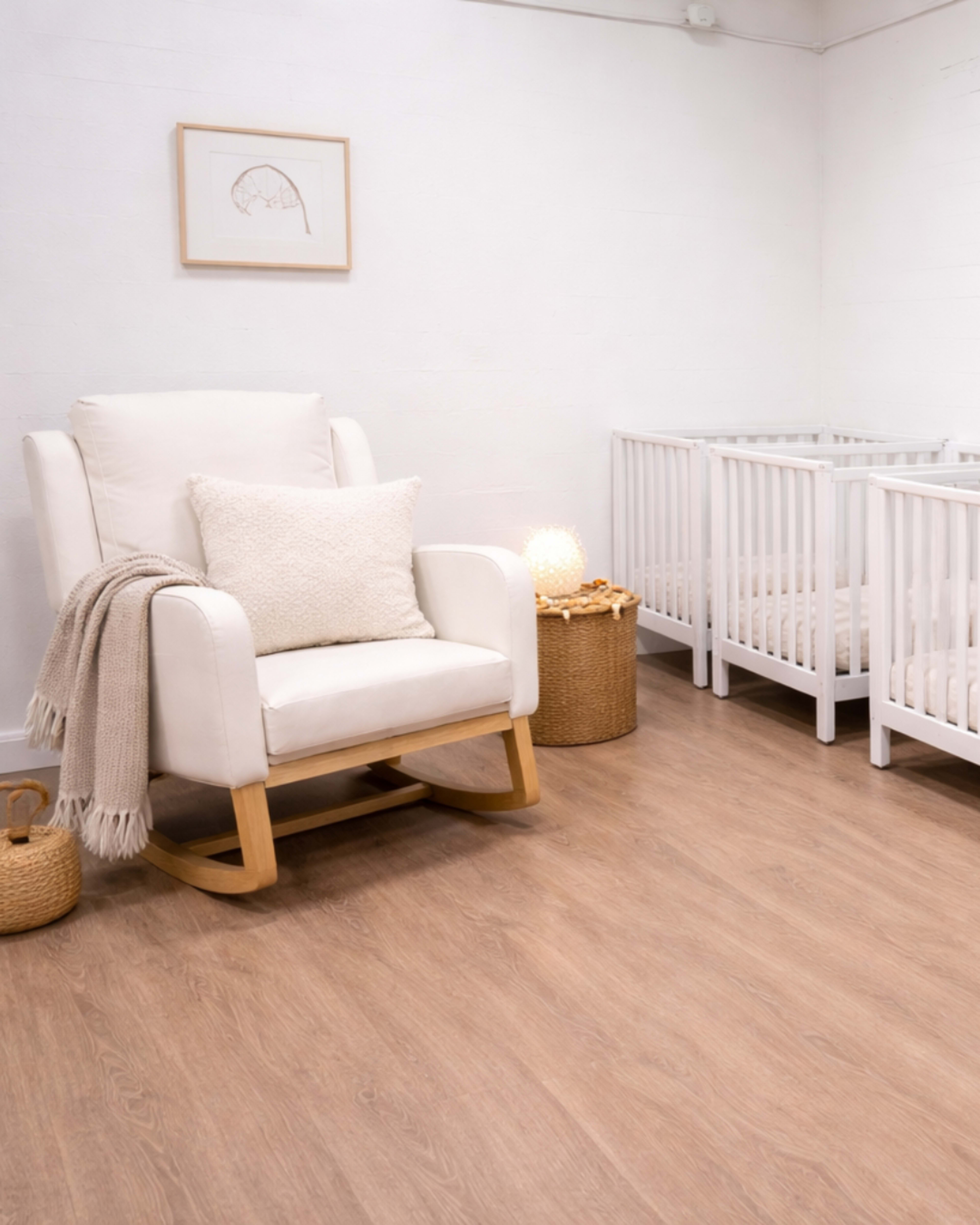 The image shows a bright nursery with a white rocking chair, a decorative pillow, and two white cribs against a light-colored wall.