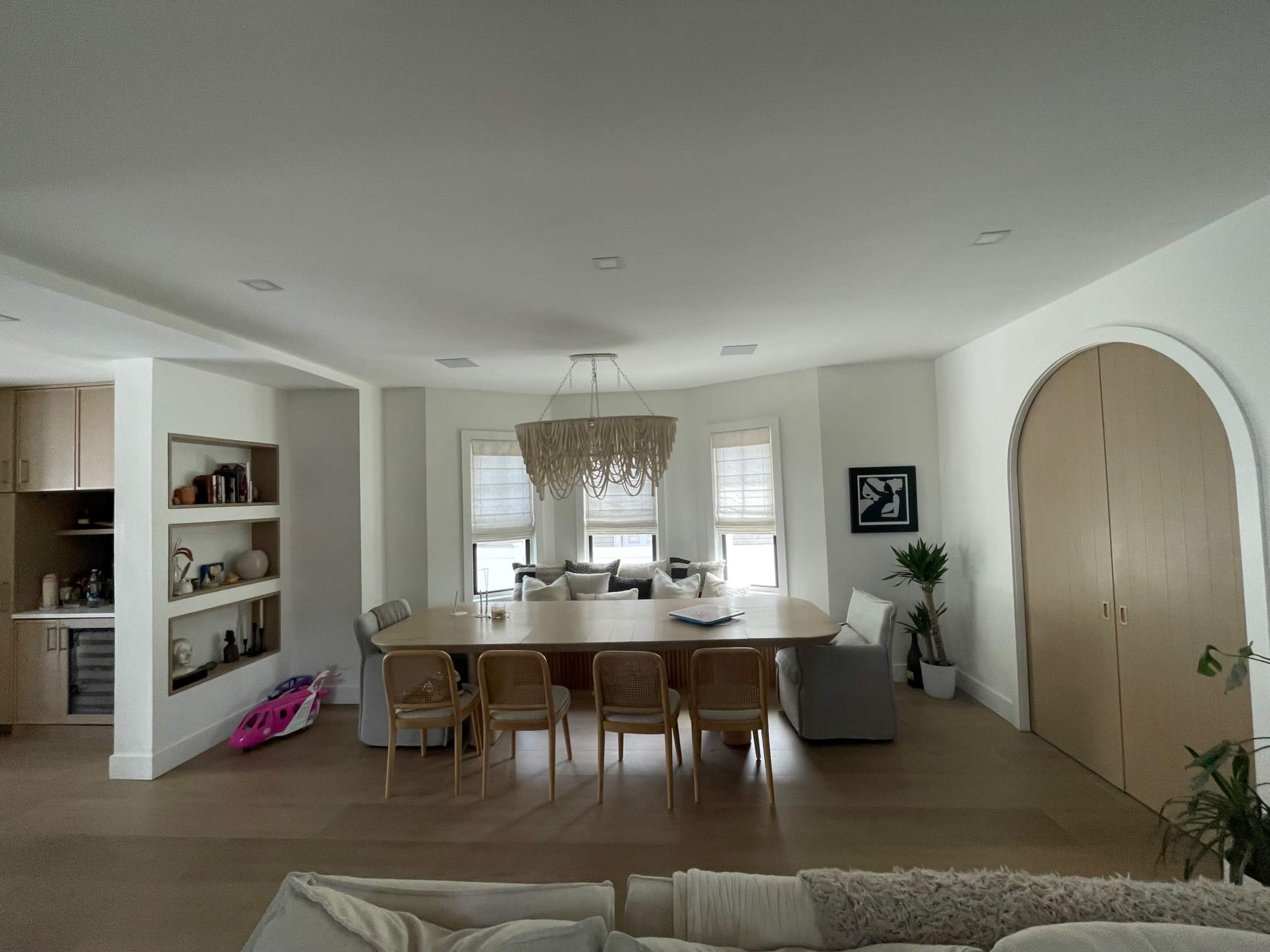 The image shows a modern dining area featuring a large table surrounded by chairs, with a light fixture above, and a cozy seating arrangement near the windows.