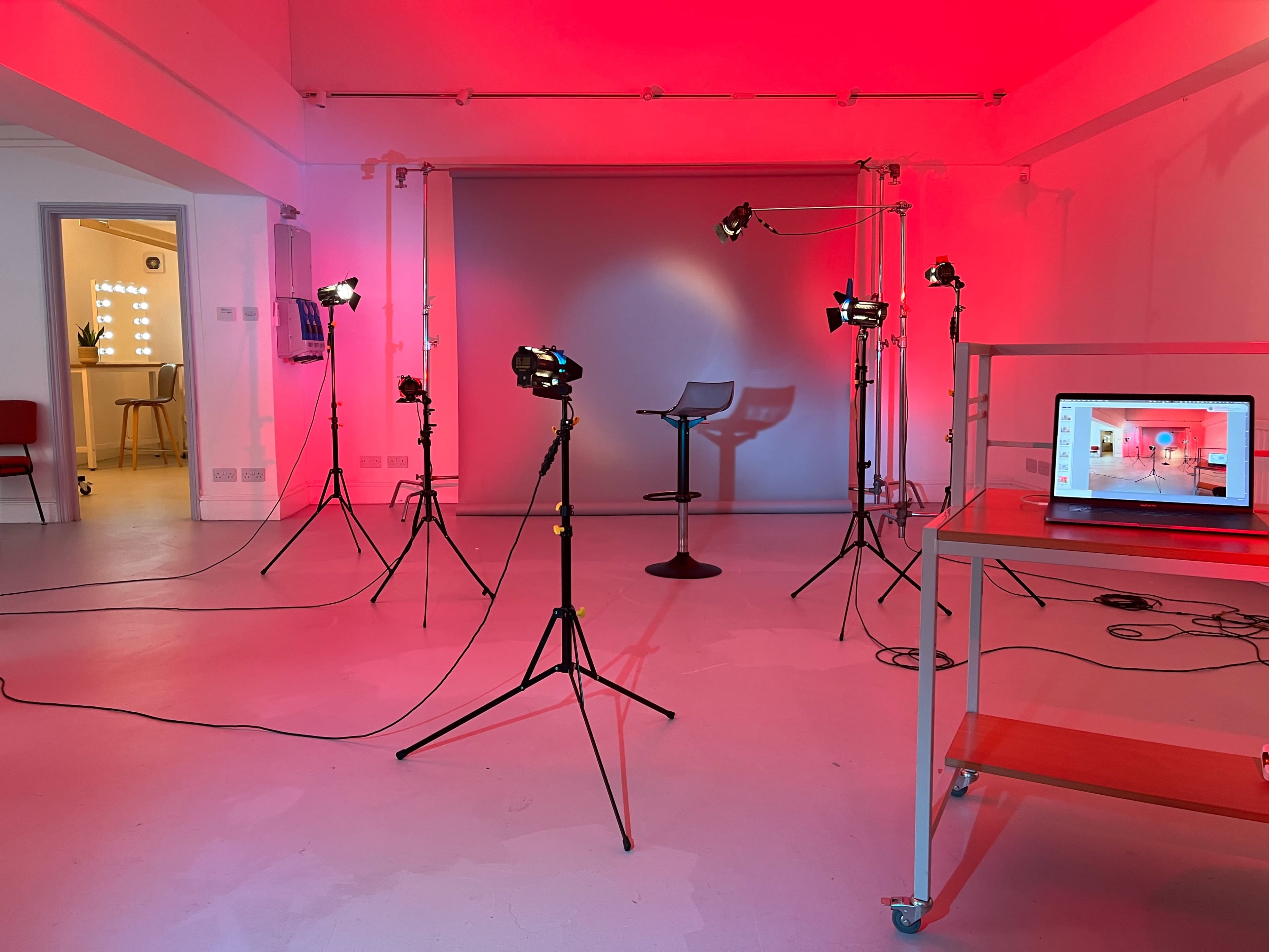 A studio space with red lighting, equipped with various photo lights on tripods, a stool, and a laptop on a table.