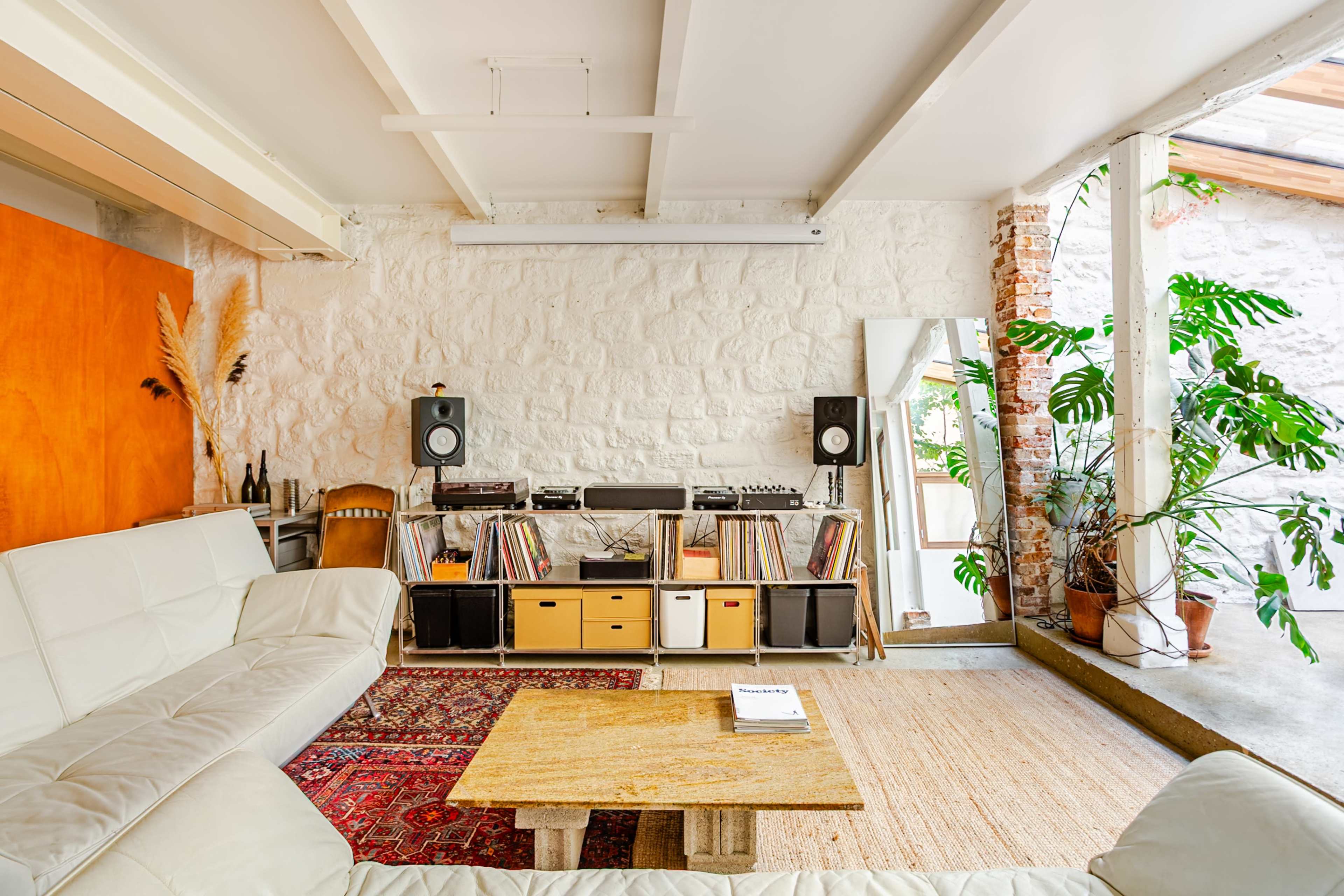 The image features a modern living room with a white stone wall, a low coffee table, a vinyl record setup, and large plants beside a bright window.