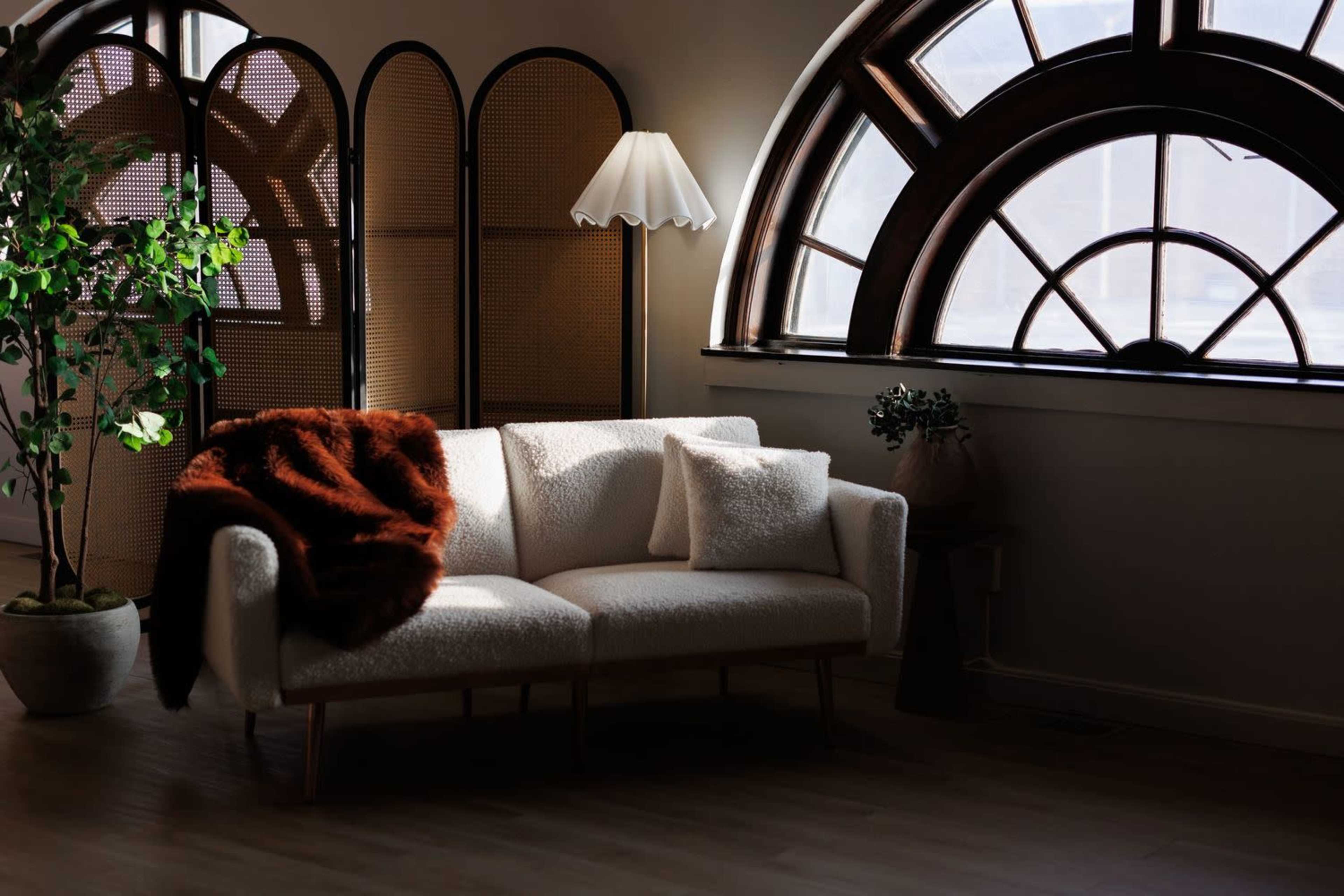 A white couch with a brown throw blanket and pillows is positioned near a large arched window, with a standing lamp and a potted plant nearby.