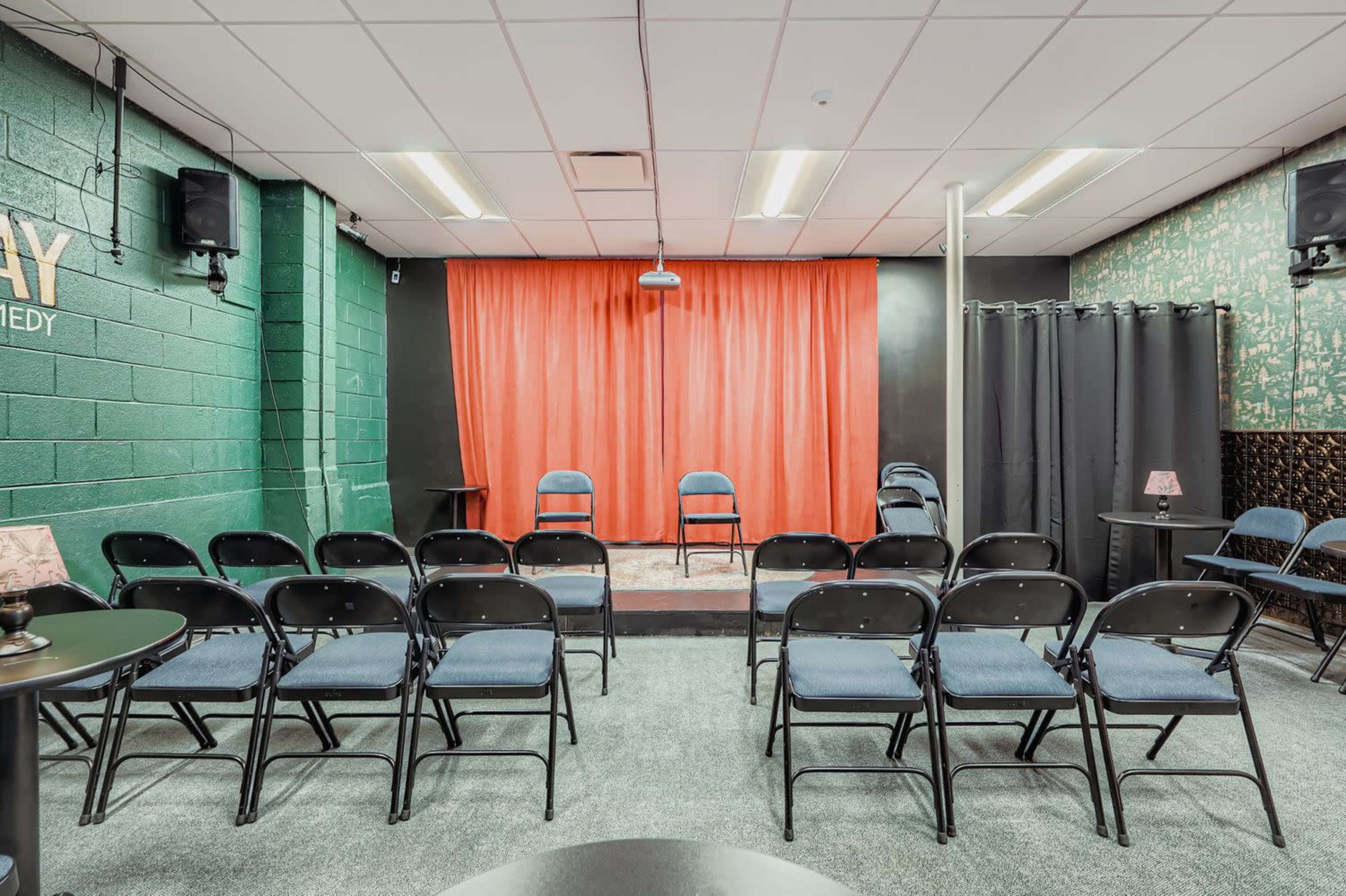 A small comedy venue features a stage with red curtains, two chairs on a platform, and rows of black folding chairs for an audience.