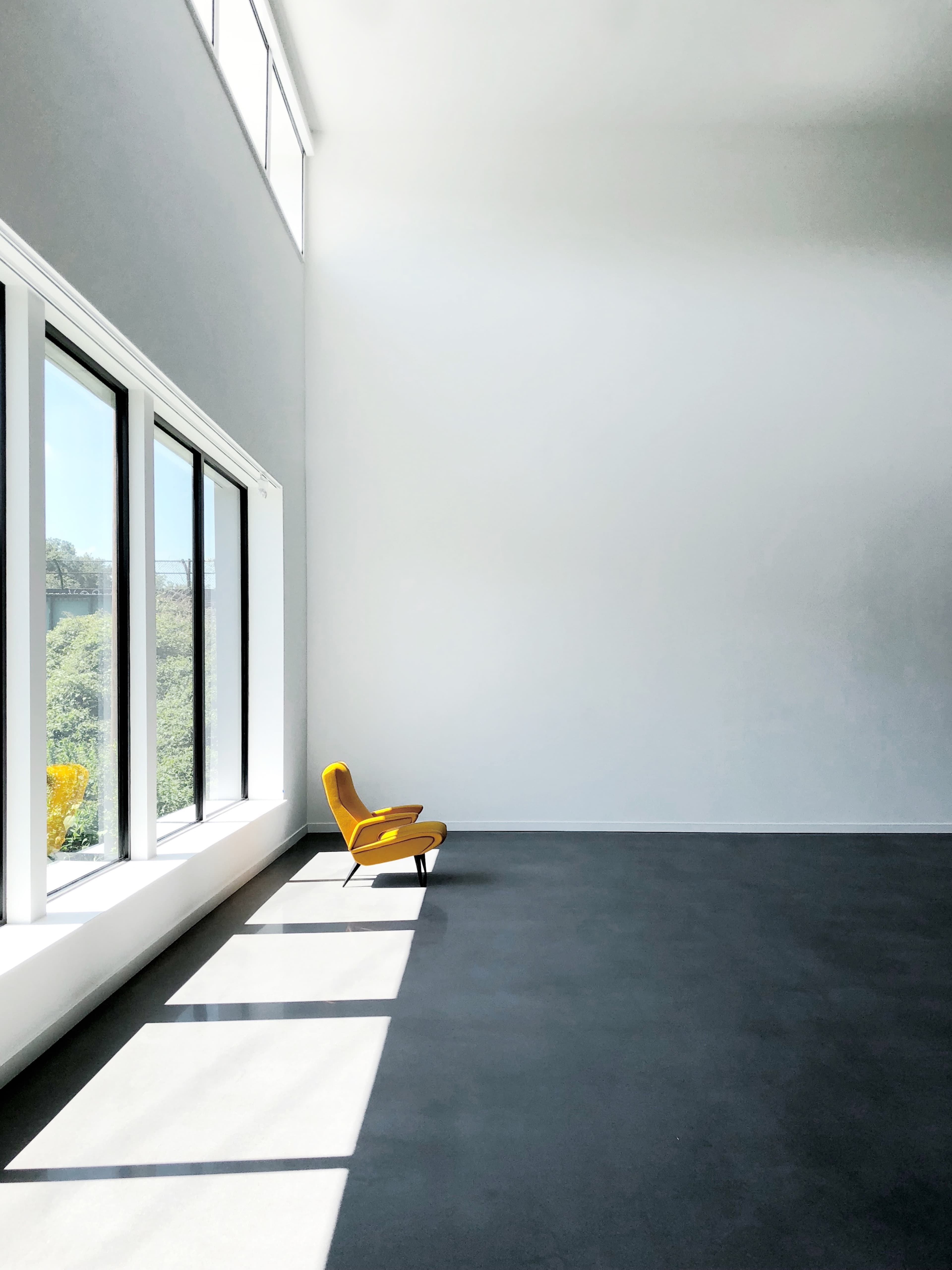 A single yellow chair is placed against a large, white wall illuminated by sunlight streaming through floor-to-ceiling windows.