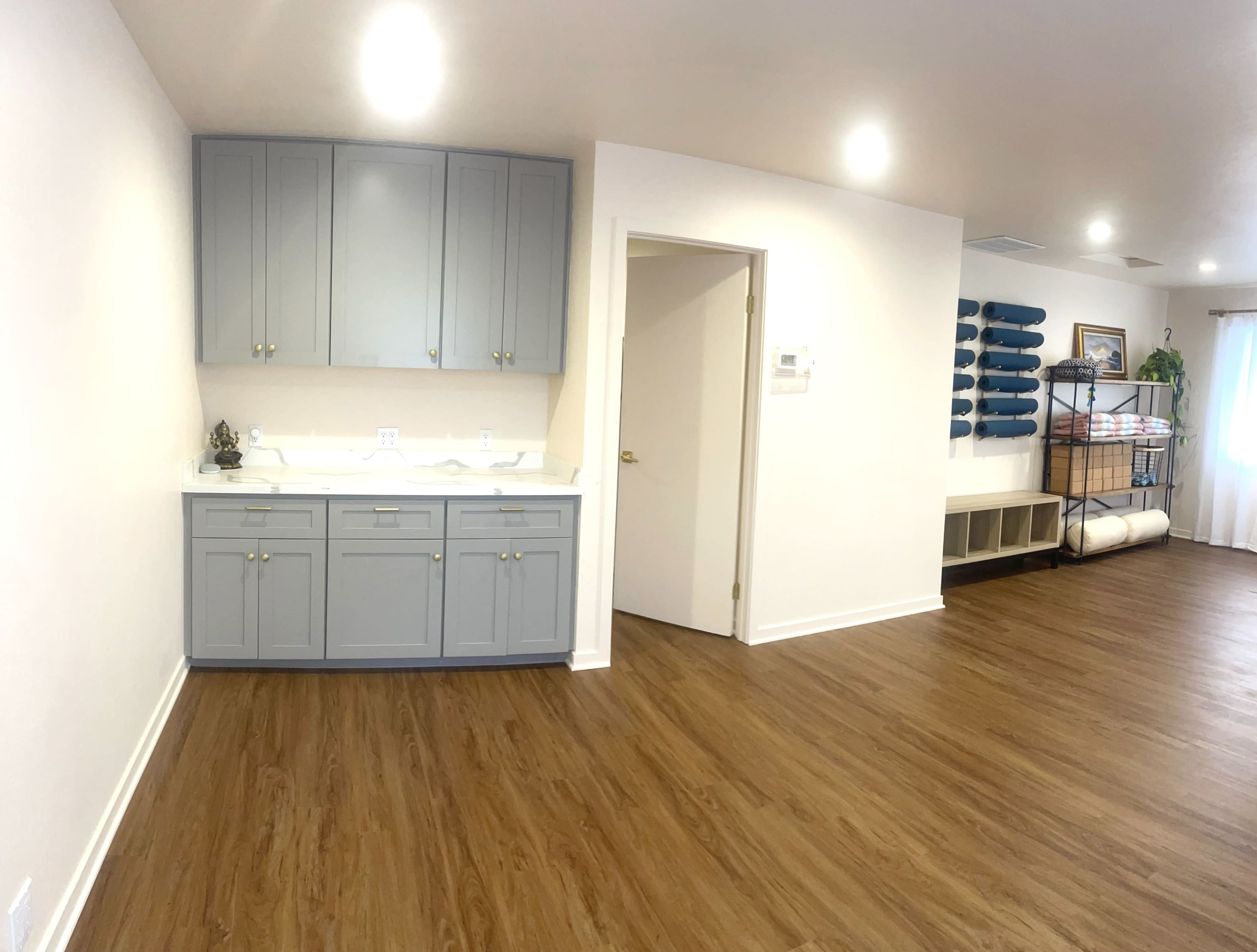 A spacious room features light-colored walls, gray cabinets, and wooden flooring, with shelves and rolled items visible in the background.