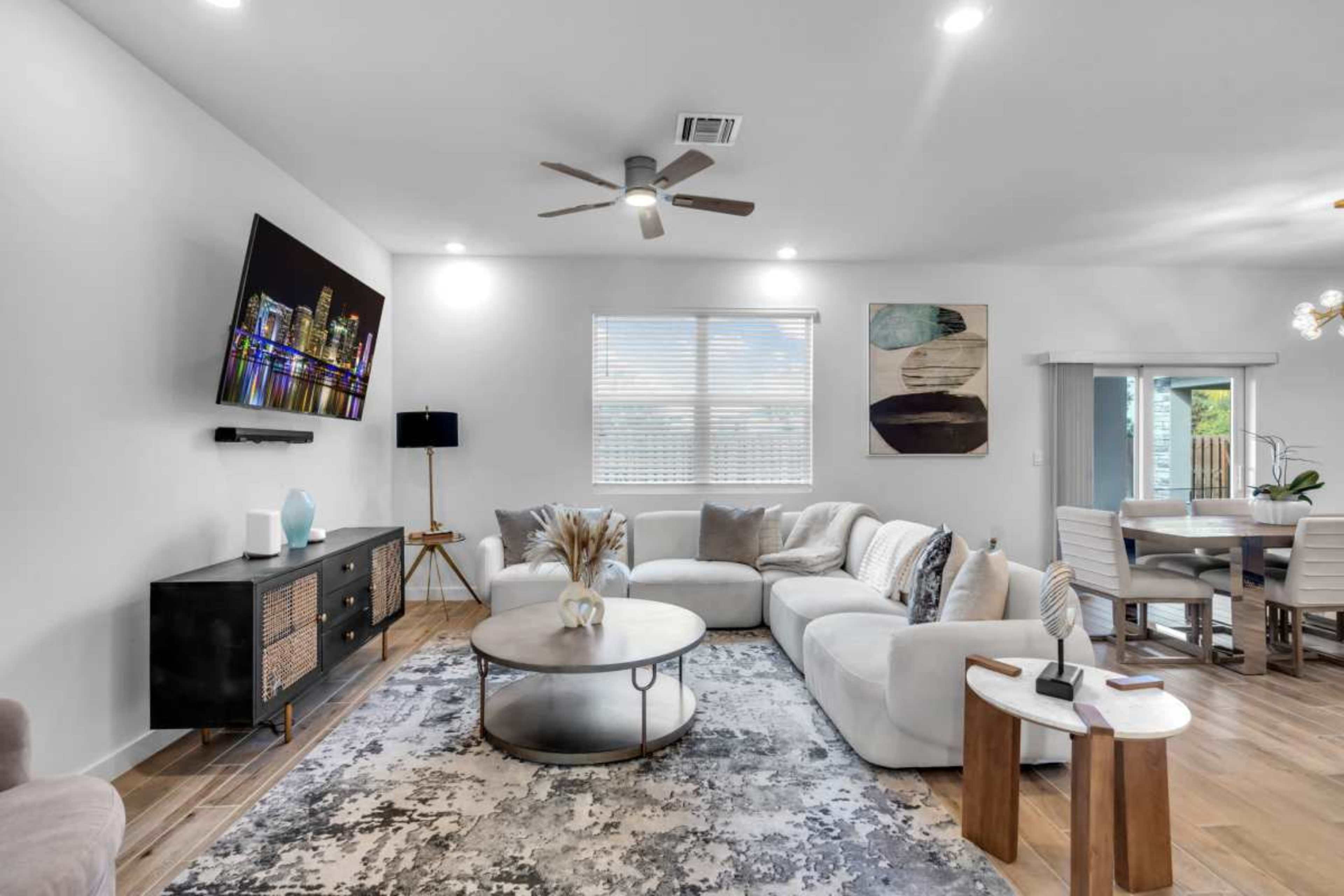 A bright living room features a large sectional sofa, a round coffee table, a wall-mounted television, and modern decor elements.