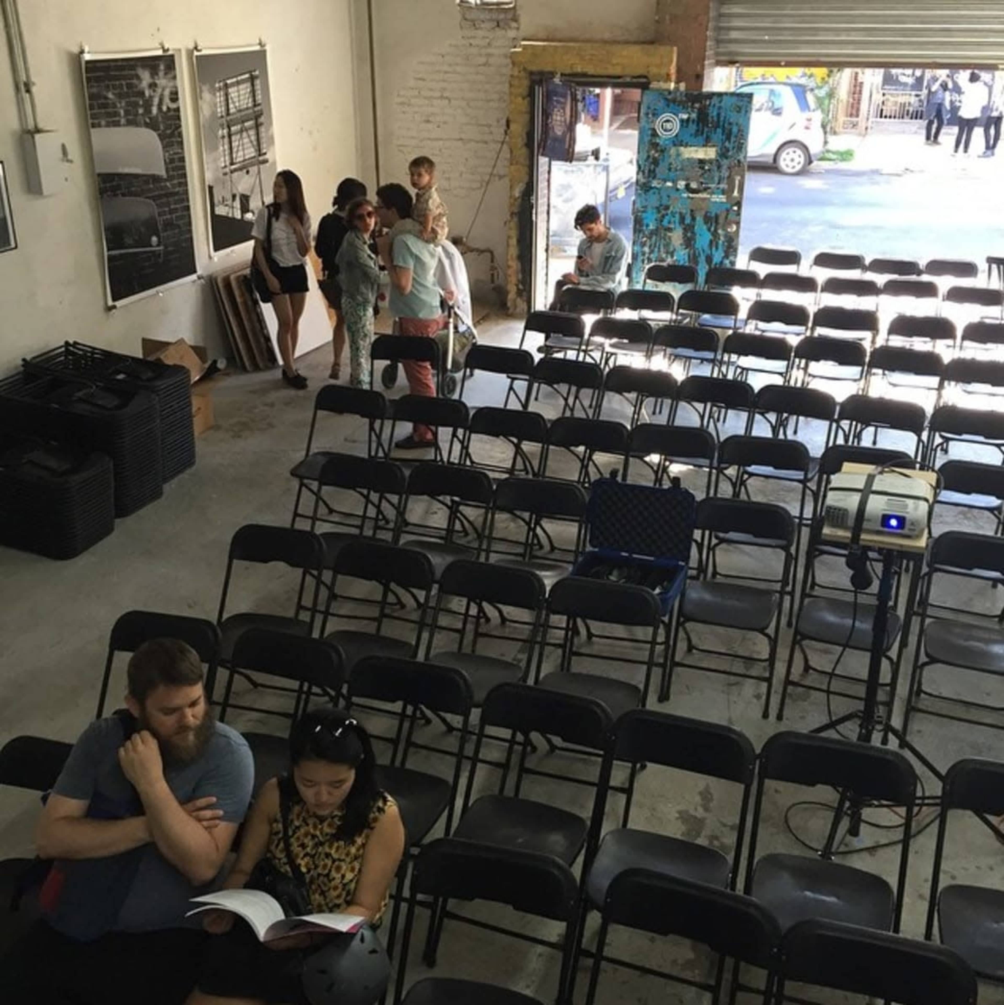 The image shows a large, empty room with rows of black chairs set up facing a wall, and a group of people scattered throughout the space.