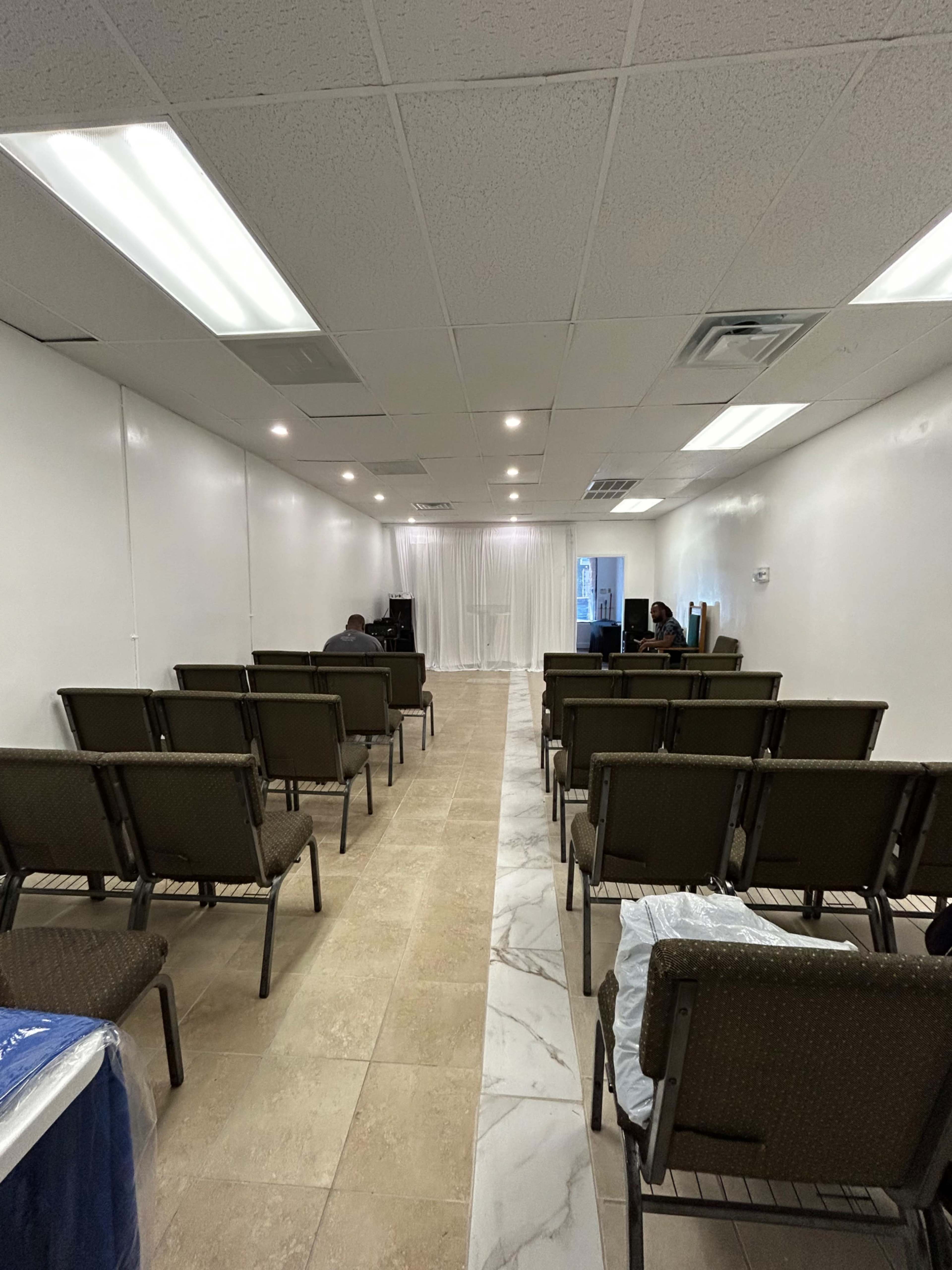 The image shows a room with rows of empty chairs facing a light-colored backdrop, suggesting a setting for a gathering or event.