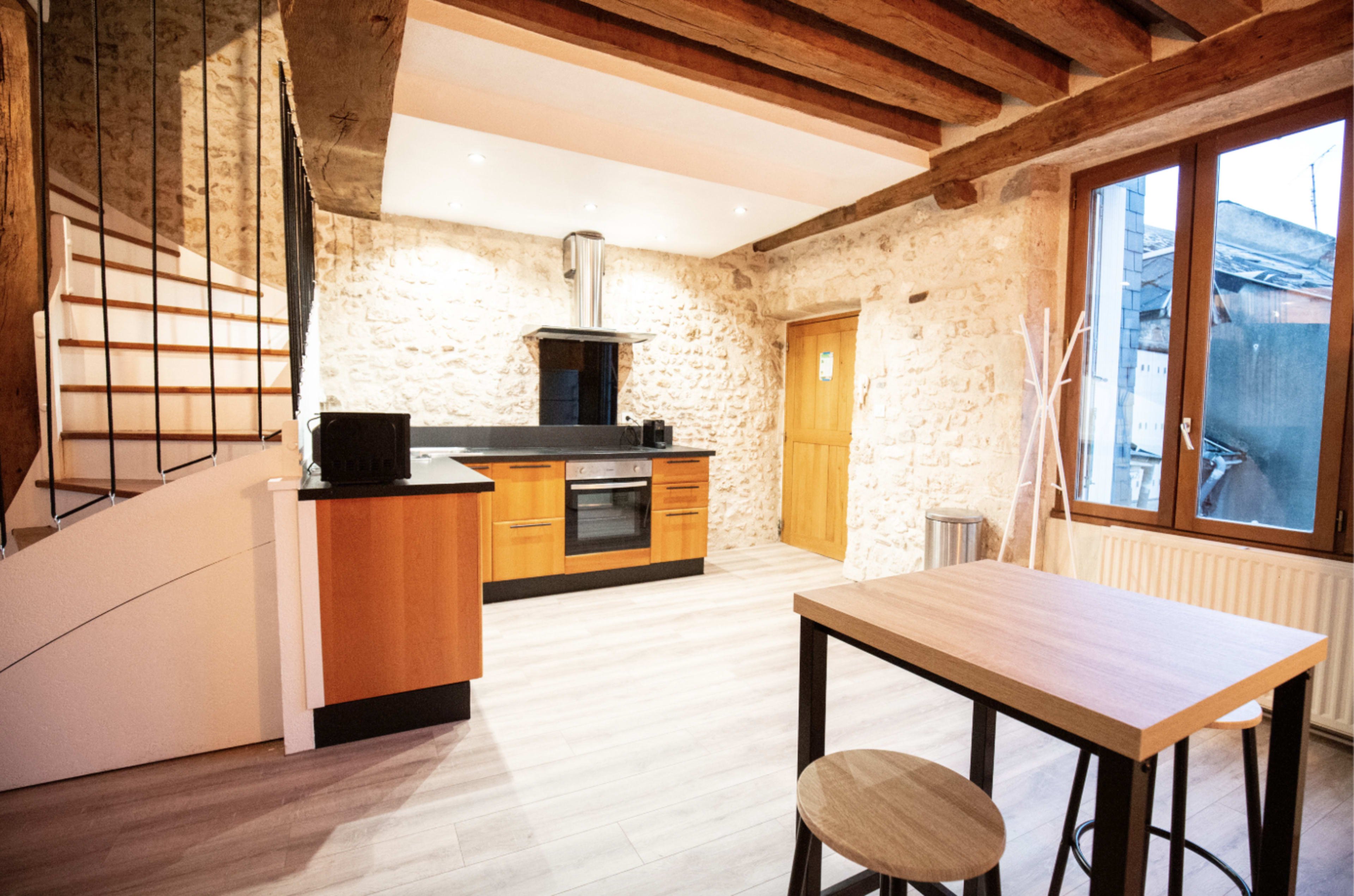 The image shows a modern kitchen with wooden beams, a staircase, and a dining table in a well-lit space featuring stone walls and a large window.