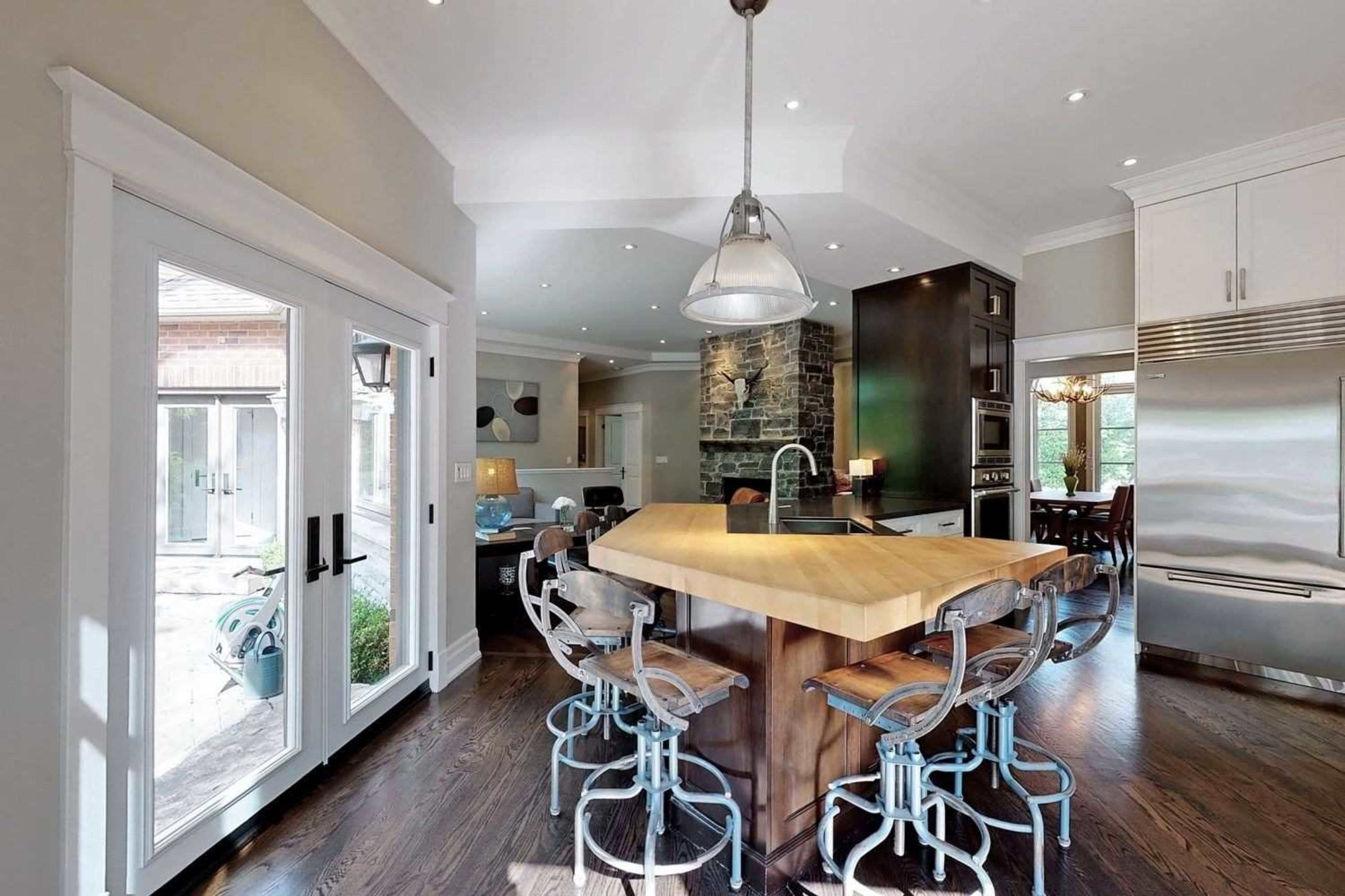 The image shows a modern kitchen featuring a large wooden island with seating, stainless steel appliances, and large windows that allow natural light to enter.