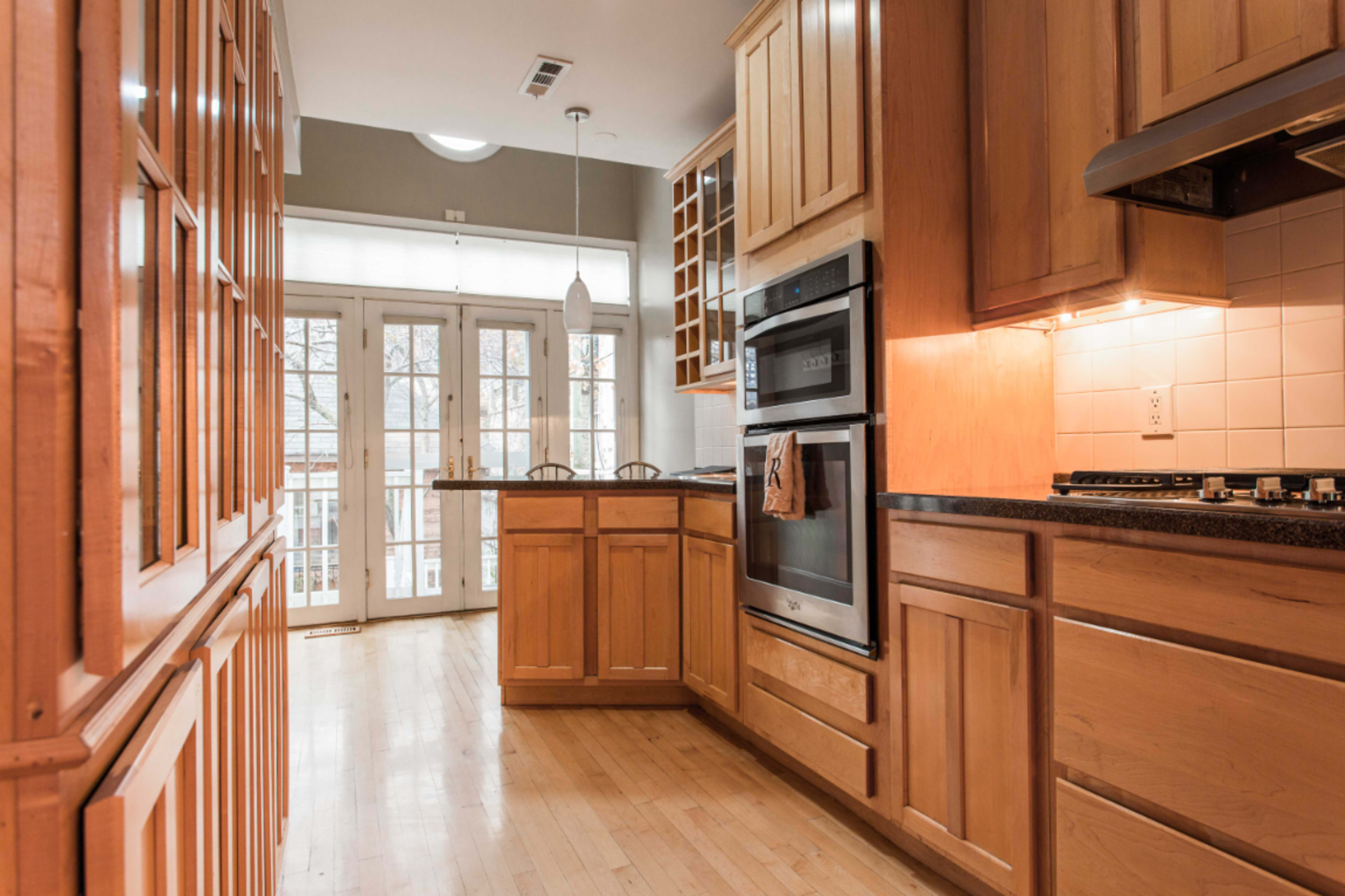 A narrow kitchen features wooden cabinets, a double oven, and a view of a patio through glass doors at the end of the room.