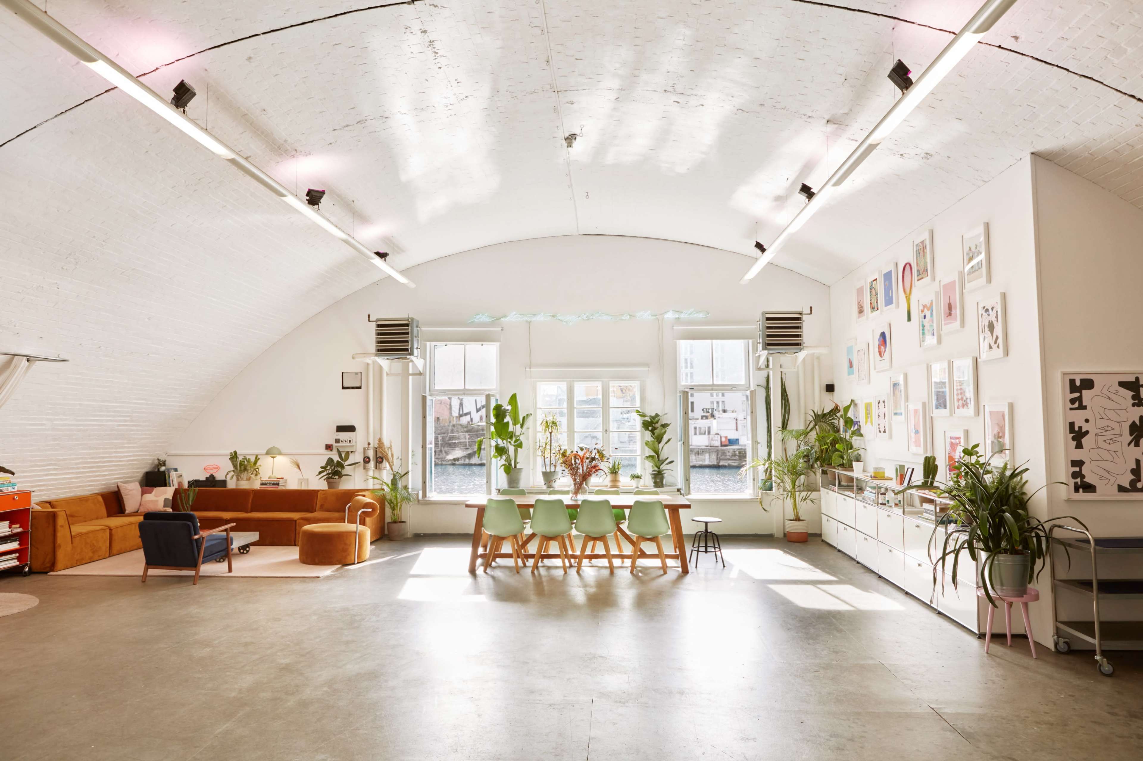 A spacious, well-lit room with a large dining table, a cozy seating area, and various plants and artwork on the walls.