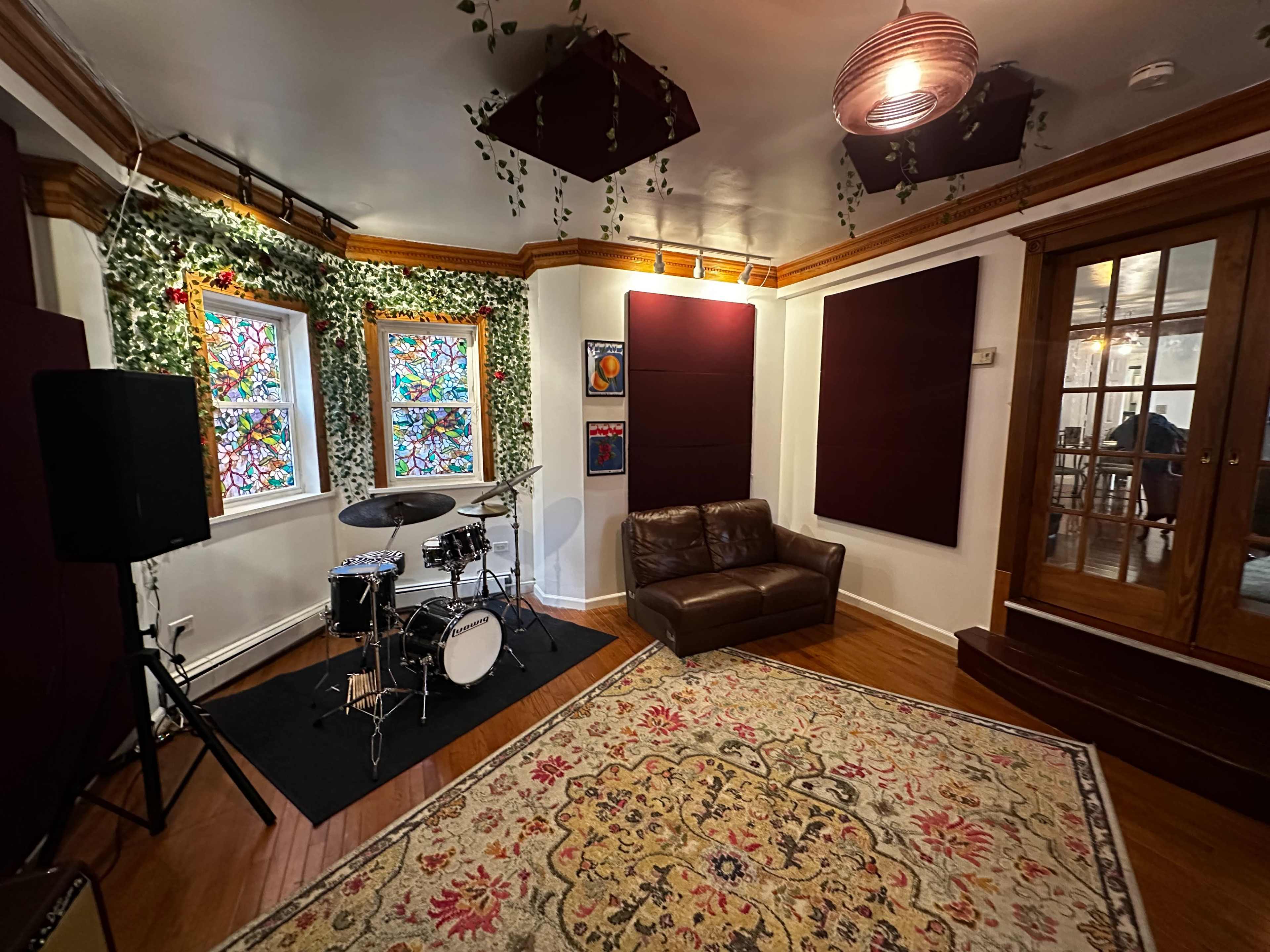 The image shows a music studio featuring a drum set, a brown leather couch, colorful stained glass windows, and acoustic panels on the walls.