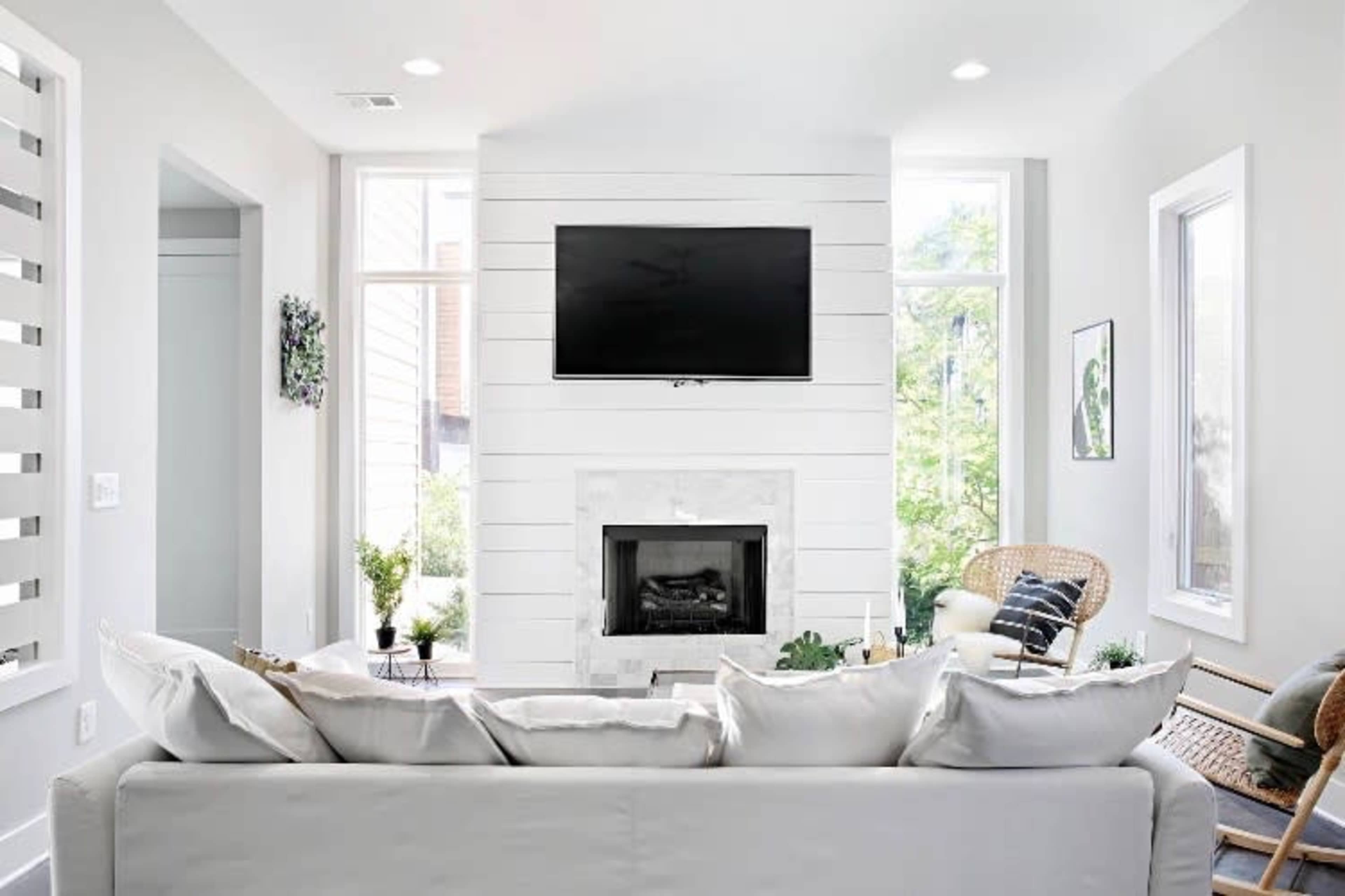 A modern living room features a wall-mounted television above a marble-accented fireplace, with large windows flanking the space and a neutral color palette.