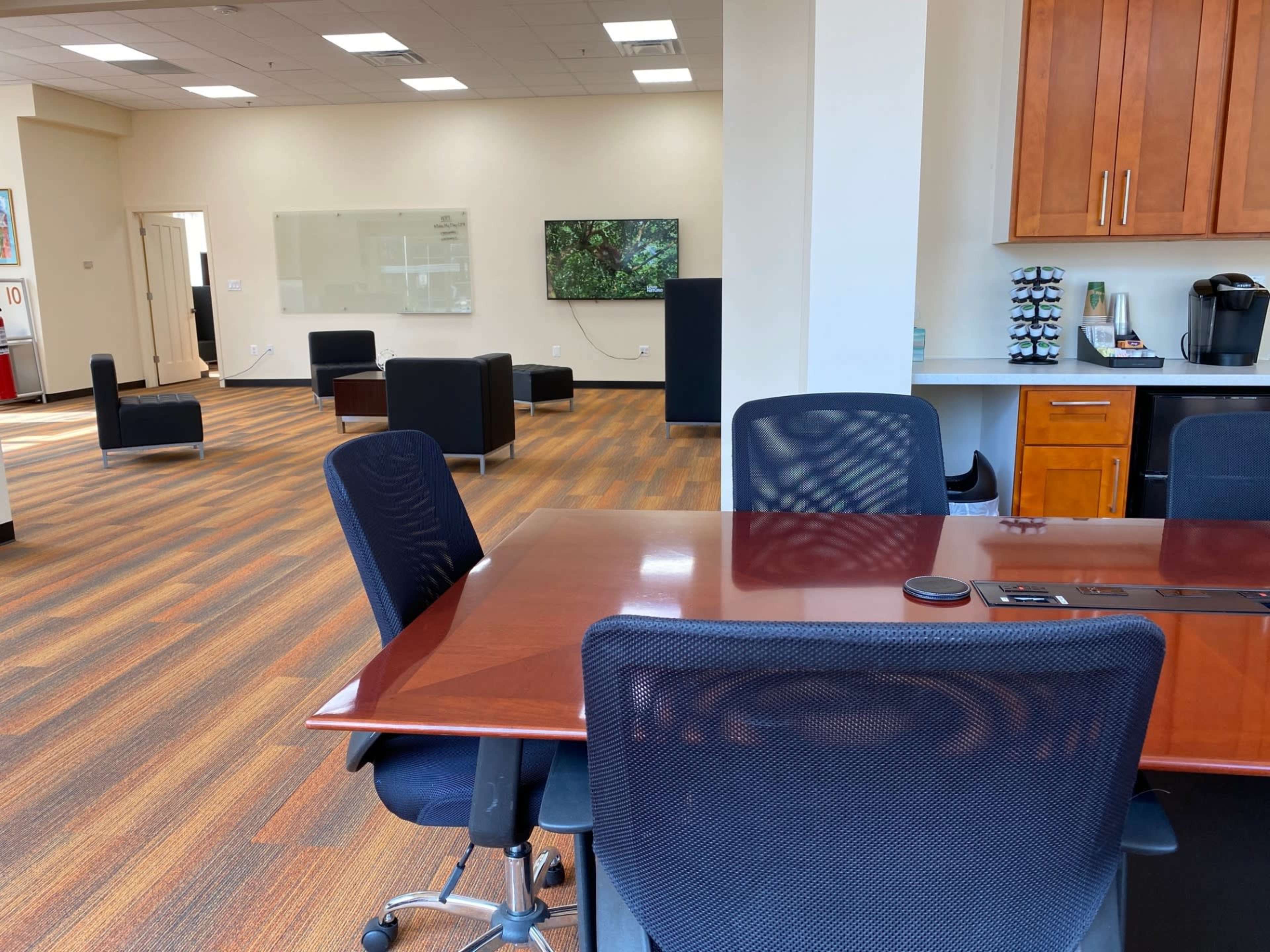 The image shows a modern office space featuring a conference table with chairs, a kitchen area with a coffee maker, and a lounge area with black seating.